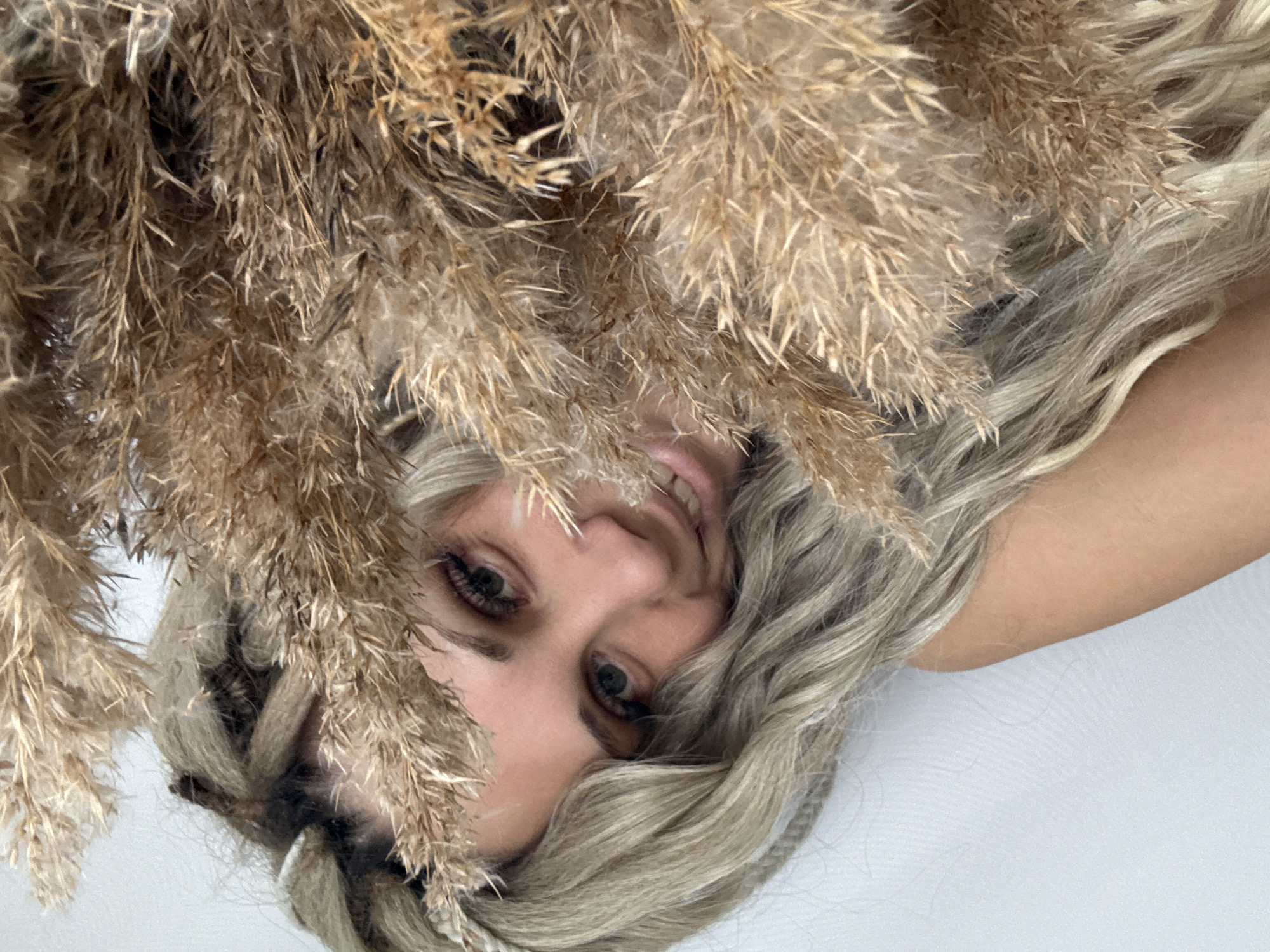 A person with blond hair, styled with dreadlocks, is positioned in front of a bundle of dried pampas grass, with their face visible through the foliage. The person is smiling slightly. The background is a light, possibly white surface. The image is taken from an unusual angle. There is no indication this photo was taken in Rusanovo, Russia.