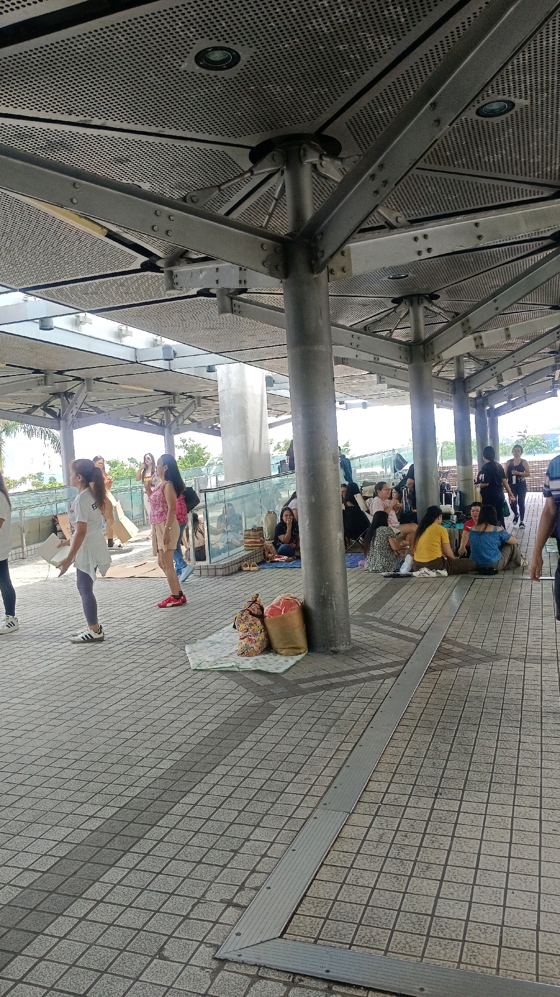 The image depicts an outdoor public space in Hong Kong, China, likely a promenade or plaza covered by a modern, perforated metal canopy supported by large pillars. The ground is paved with light-colored square tiles. In the foreground, there are two large bags placed on a mat on the tiled surface.
The scene features a group of people, predominantly women, gathered in a shaded area. Several individuals are seated on the ground on mats or blankets, some appearing to be resting or socializing. There are also a few people standing. In the middle ground, a woman in a white top and skirt is walking, and another woman in a pink top and shorts is standing. Further back, a group of people are seen near a set of escalators or a sloped walkway. The environment suggests a moderately busy day with people utilizing the outdoor space.