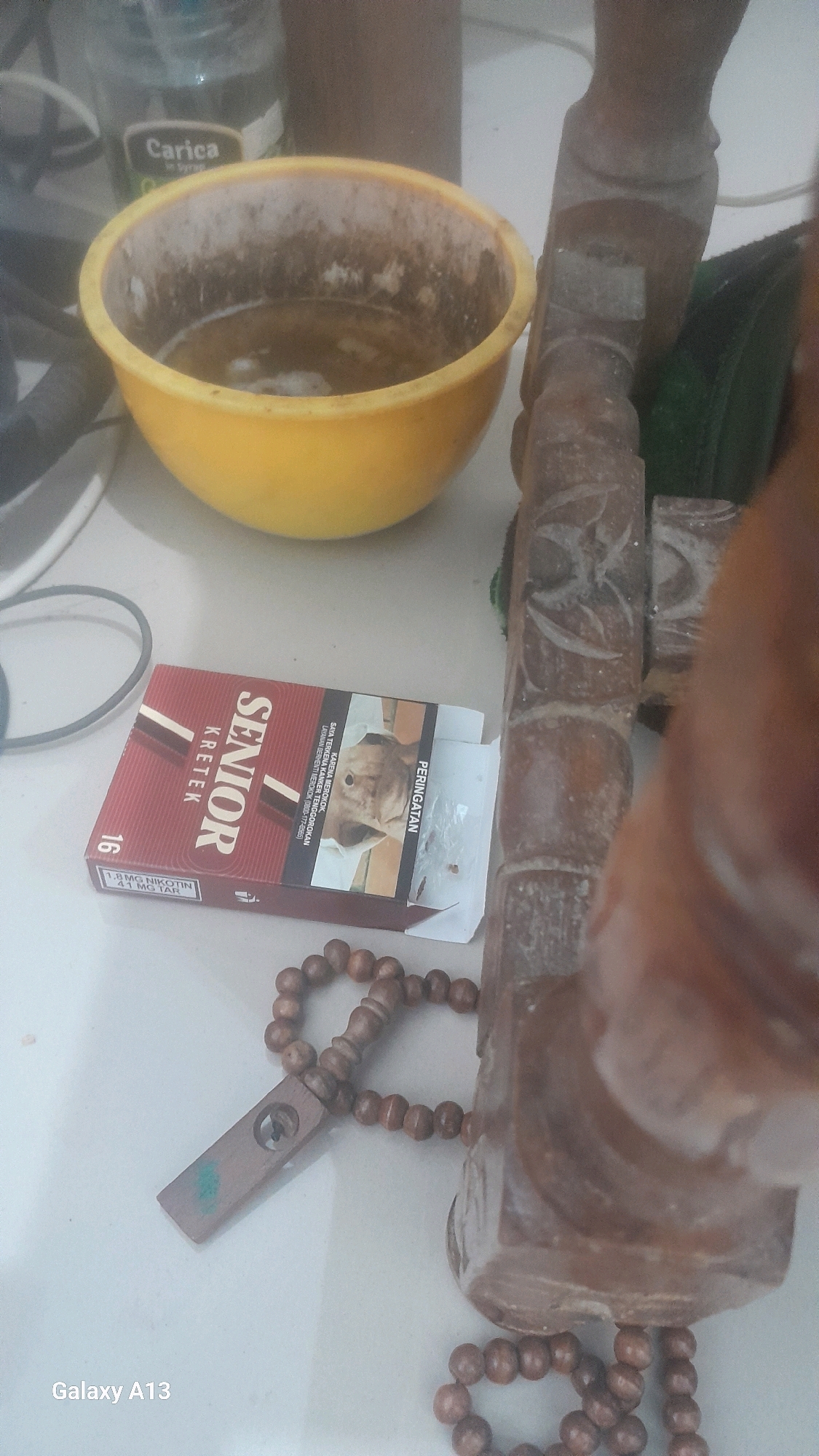 The image shows a still life scene on a white surface, possibly a tabletop. In the foreground, a pack of "Senior Kretek" cigarettes is partially open, revealing a warning label with an image of a dog. Next to the cigarette pack, a string of wooden prayer beads with a small wooden pendant lies on the surface. To the left of the cigarettes is a yellow bowl containing a murky, light-brown liquid with some white residue on the inner rim. Behind the bowl, a partially visible bottle with a label reading "Carica" can be seen. To the right of the cigarettes and prayer beads, a section of a carved wooden object, possibly an idol or a decorative piece, is prominent, appearing to be made of dark wood with intricate carvings. The lighting is dim, and the overall scene suggests a collection of personal items. The location is not discernible from the image itself, and therefore, its connection to Hadipolo, Indonesia, cannot be confirmed or described.