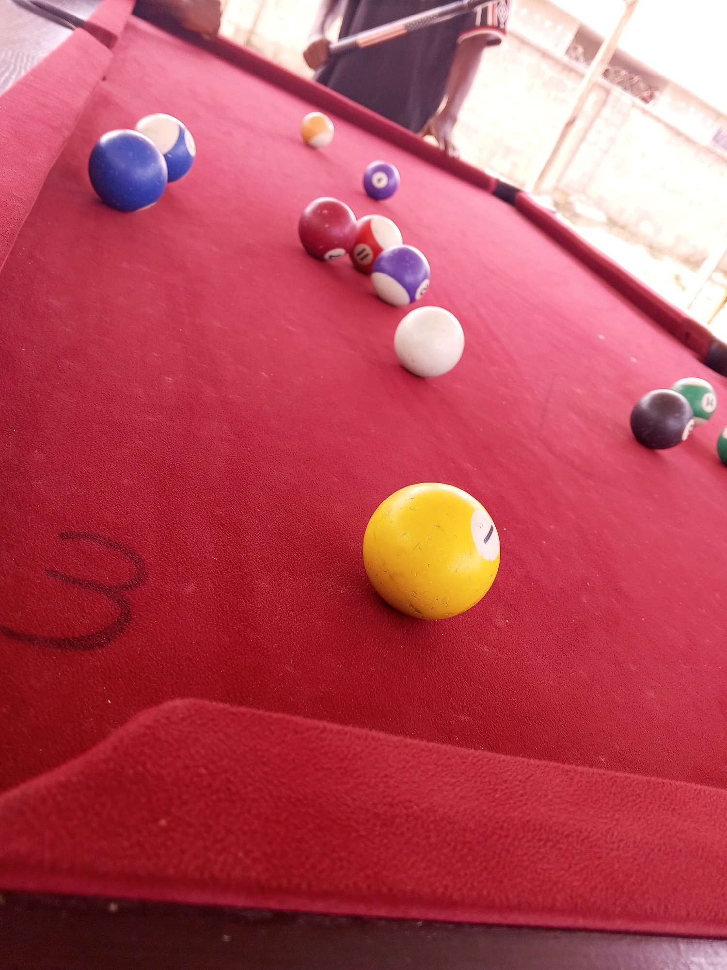 This media file shows a close-up of a red pool table with several billiard balls scattered across it. The perspective is low, looking across the table towards a person in the background. The person, only partially visible, appears to be playing pool, with a cue stick held in their hands.
The billiard balls include a solid yellow ball with the number "1" visible, a white cue ball, several red balls, some purple and white striped balls, and a dark brown ball with some green markings. The pool table surface is a vibrant red felt, and the edge of the table is also visible. The background is out of focus, but suggests an indoor or covered outdoor setting with natural light. The overall impression is that of a casual game of pool taking place.
There is some text visible on the person's shirt in the background, which appears to be "TIR" or something similar. The lighting suggests it is daytime. The location is identified as Lokoja, Nigeria, which may imply a typical leisure activity in that region. No specific emotions are discernible due to the focus and framing of the image.