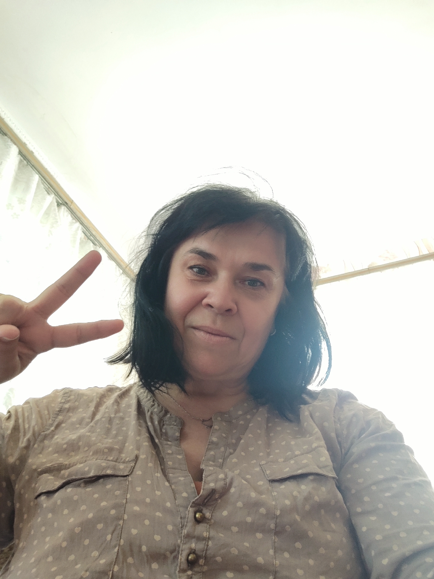 This is a selfie of a woman with dark, shoulder-length hair. She is wearing a light brown, polka-dotted button-up shirt. She is looking at the camera and smiling, holding up two fingers in a peace sign with her left hand. The lighting is bright, suggesting it is daytime, and the background appears to be an indoor setting with white curtains or blinds. There is no visible text in the image. The woman's expression appears happy and relaxed.