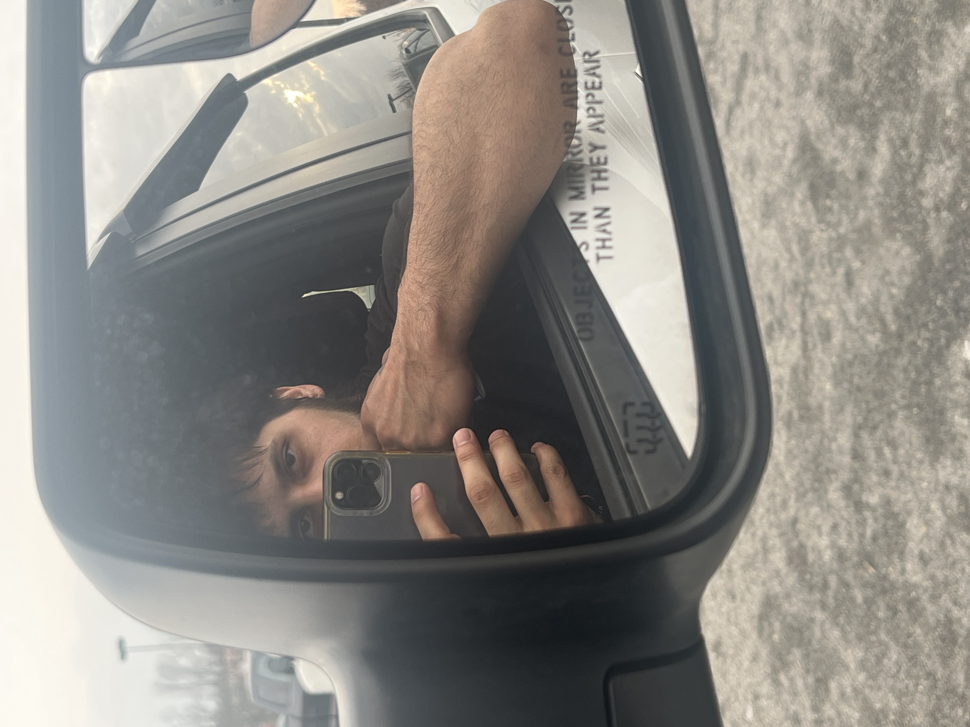 A person with dark hair is taking a picture of themselves in a side view mirror of a vehicle. The mirror reads "OBJECTS IN MIRROR ARE CLOSER THAN THEY APPEAR". Outside the mirror, a gray surface is visible, along with other vehicles in the background. The exact location in Orange Lake, United States, cannot be determined from the image.