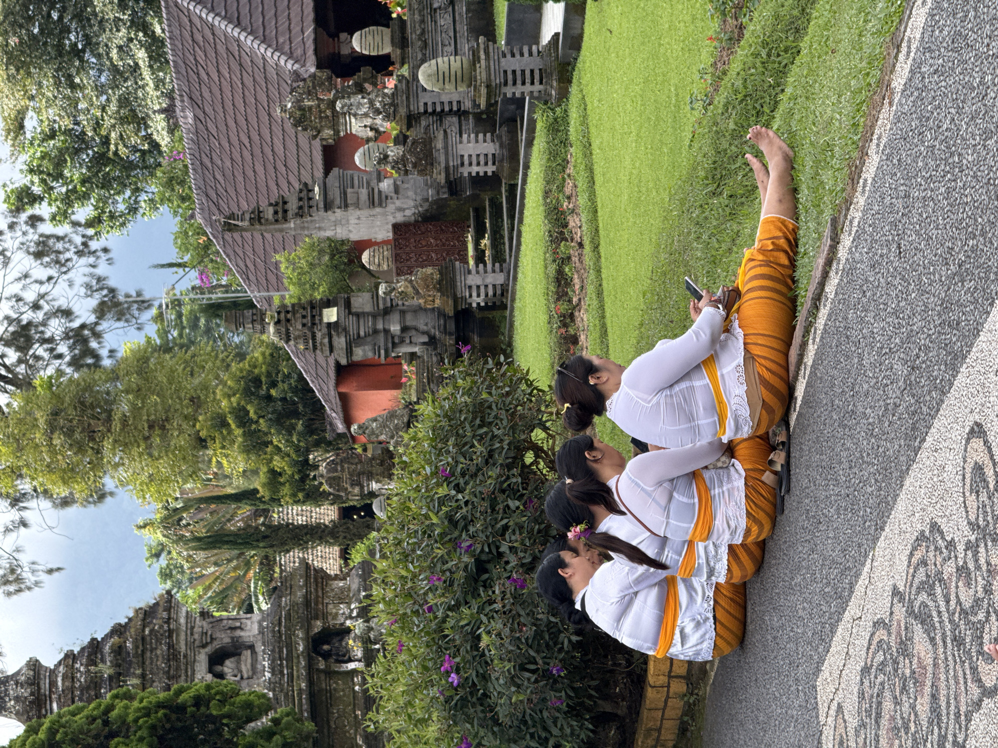 The image depicts three women seated in a row along a patterned stone pathway, adjacent to a lush green lawn. They are barefoot, wearing traditional Balinese attire consisting of white blouses, orange and brown striped sarongs, and yellow sashes. One of the women is using a mobile phone. Behind them is a densely vegetated area with shrubs and flowering plants, in front of traditional Balinese architecture. The architecture includes a building with a tiled roof and intricately carved stone structures, indicating a temple or cultural site. The environment consists of tropical greenery, including trees and manicured lawns, giving the impression of a well-maintained garden or park area in Baturiti, Indonesia.