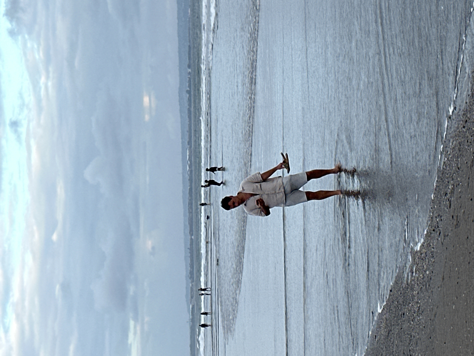 A man stands in shallow water at the beach in North Kuta, Indonesia, looking at his phone and holding a sandal in his left hand. He is wearing a light-colored linen shirt and white shorts. Several other people are in the background, some walking in the water and others in the distance. The ocean has gentle waves and a cloudy sky looms overhead. The shore is covered in dark sand and small pebbles.