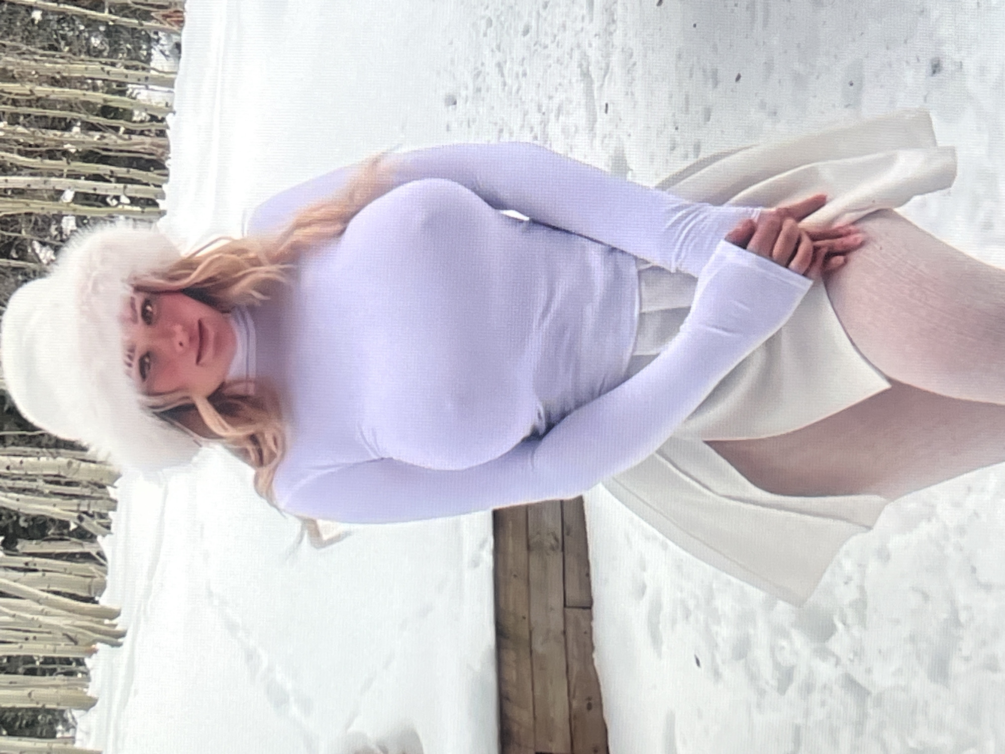 A young woman stands in a snowy landscape, wearing a white turtleneck long-sleeved shirt, white fingerless gloves, white tights, and a fluffy white hat. Her blonde hair is visible around her face. She is looking towards the camera with a slight smile. Behind her, a wooden structure is partially visible, and the background consists of snow-covered ground with some bare trees. The scene appears to be set during the daytime, likely in a cold, winter environment given the snow. There is no visible text in the image.