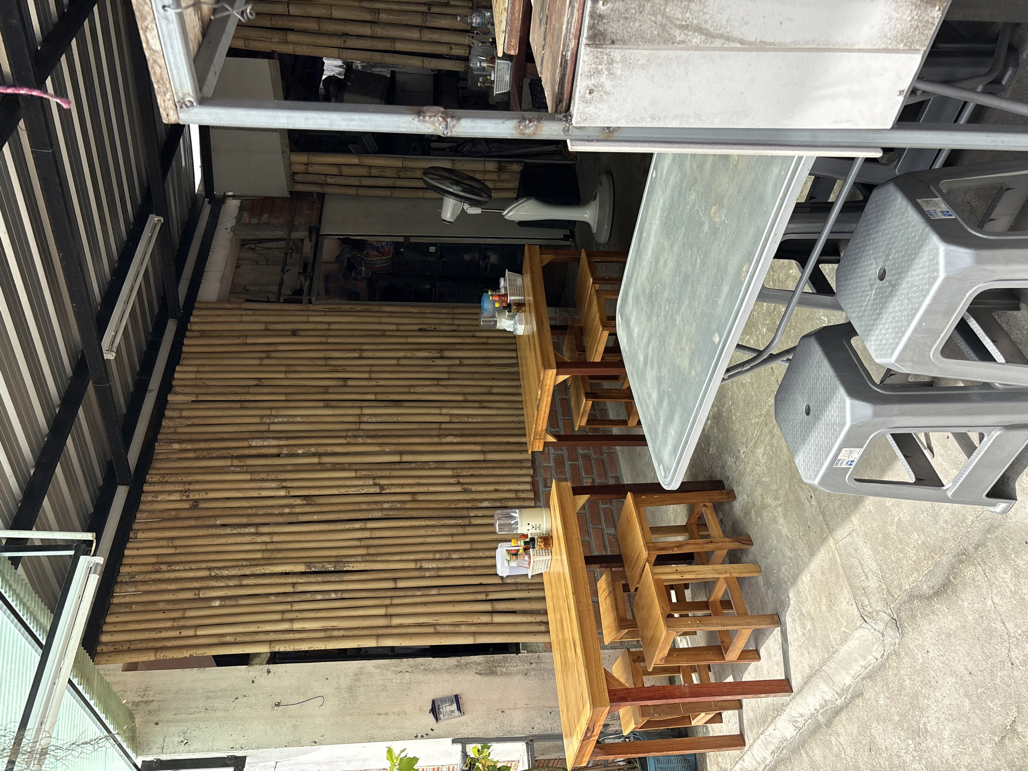 This is a medium-shot, eye-level, outdoor photograph of a food stall or small restaurant in Choeng Thale, Thailand. The scene is set under a corrugated metal roof supported by black metal beams. The walls are made of a rough concrete-like material on the left and vertical bamboo poles for the rest of the visible structure.
In the foreground, a large, rectangular, gray metal table with a textured surface is positioned. Beneath this table, two gray plastic stools with square seats and open sides are visible.
To the left of the foreground table, a rectangular, light-wood dining table with four matching wooden stools is set up. This table has a small basket on it containing bottles of condiments and two small bowls.
Further to the right, another similar light-wood dining table is set up with two wooden stools.
In the background, a white oscillating floor fan is positioned between the tables, facing towards the right. The wall behind the tables is covered with vertical bamboo poles. A doorway or opening is visible in the bamboo wall, revealing a dimly lit interior with some stored items.
The ground is paved with concrete, showing some cracks and a raised concrete curb on the left. Sunlight casts distinct shadows across the ground. The overall impression is a simple, functional outdoor dining area.