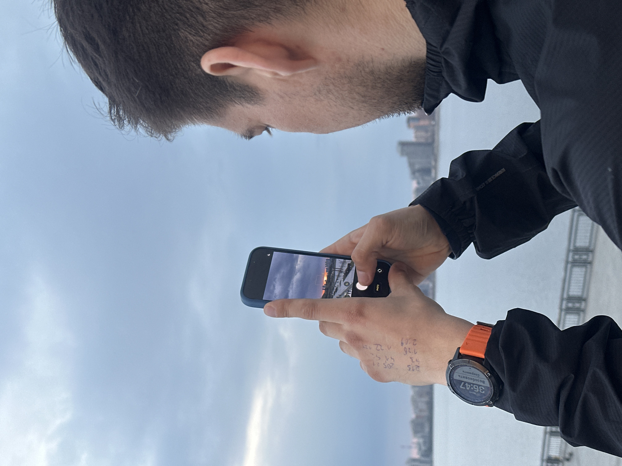 A person with short dark hair is holding a blue smartphone, taking a picture of a sunset or sunrise over a body of water. The screen of the phone displays the scene being photographed. The person is wearing a black jacket and an orange-banded smartwatch on their left wrist. Writing is visible on the person's left hand. In the background is a waterfront area with the indistinct silhouette of buildings. It is likely that this is a picture of the Kazan cityscape, Russia, given the watch displaying the Russian words "Возобновить" (Resume) and "Сохранить" (Save).