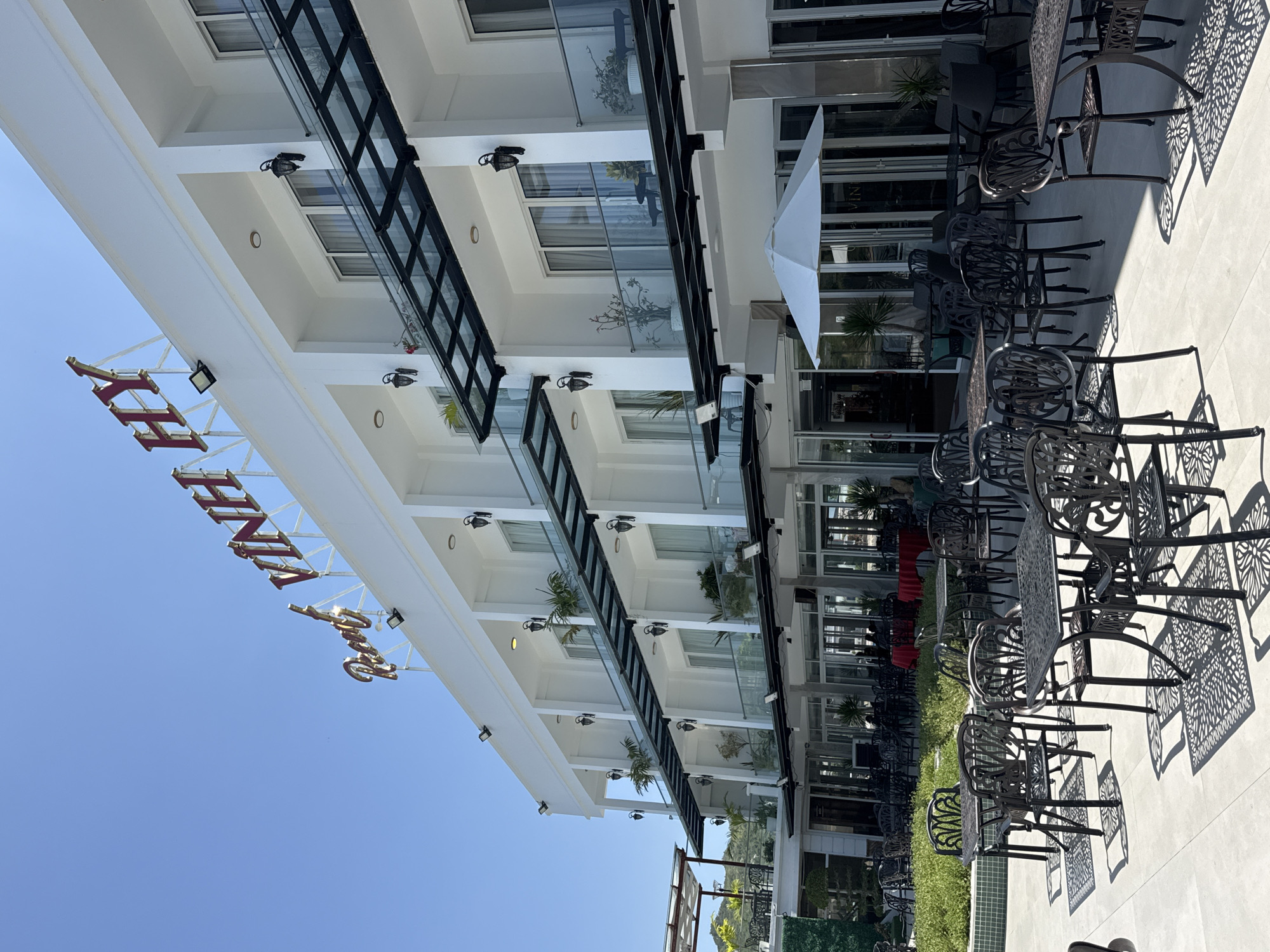 This is a photograph of the Resort Vinh Hy hotel in Vinh Hai, Vietnam. The resort features a multi-story white building with multiple balconies, each with glass railings and dark-colored outdoor lights. A prominent red sign with gold lettering spelling out "Resort Vinh Hy" is visible at the top of the building against a clear blue sky. In front of the building, an outdoor seating area is set up with many black metal tables and chairs, some with intricate patterned backs. A white umbrella is open in the center of the seating area. Some of the tables have decorative shadows cast on the paved ground from the sunlight filtering through the chairs. In the background, the resort appears to be located in a somewhat elevated area, with hints of greenery and other buildings visible in the distance.