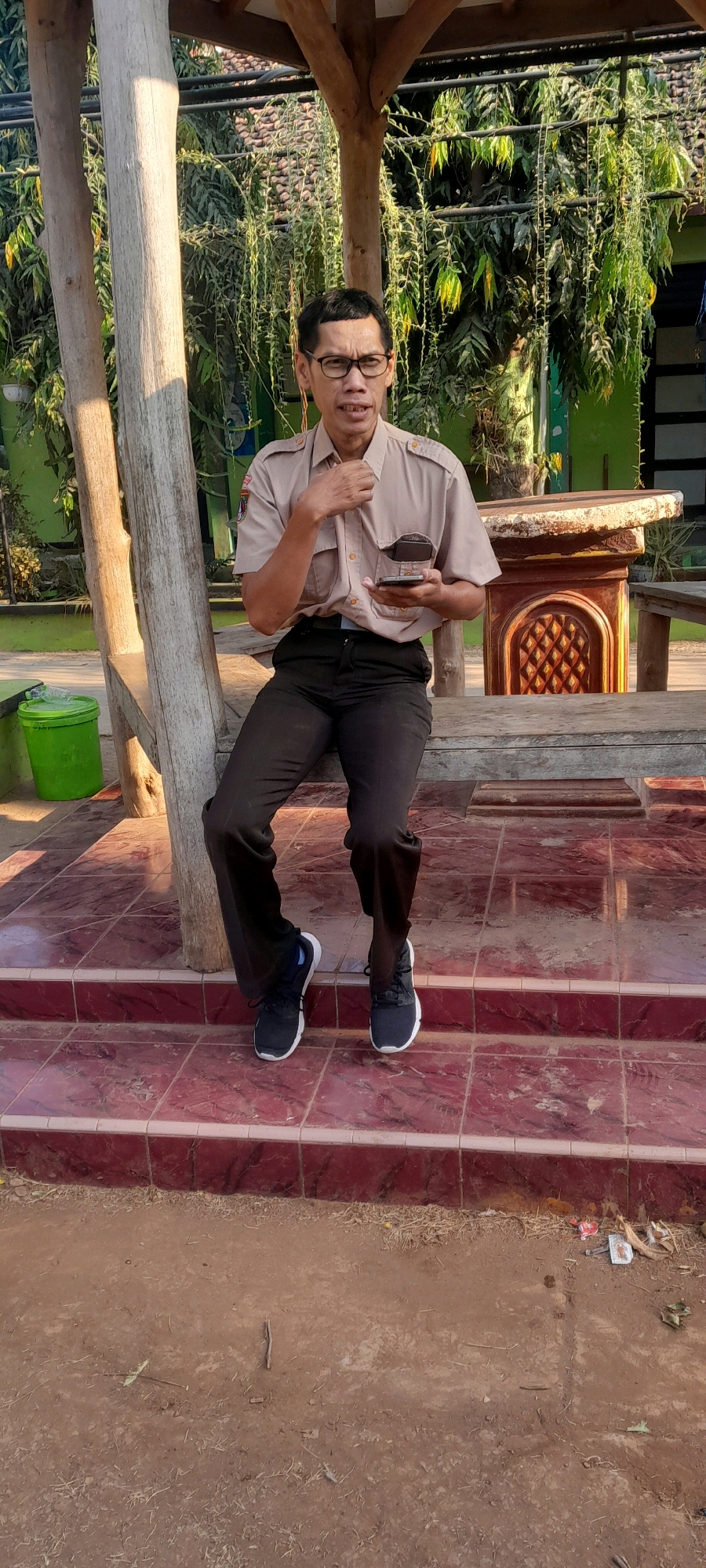 In Tahunan, Indonesia, a man wearing glasses and a light brown uniform shirt with epaulets and a patch on the left chest, and dark pants, is seated on a wooden bench. He is holding a dark-colored mobile phone in his right hand and appears to be speaking. He is wearing dark athletic shoes with white soles. The bench is positioned on a tiled step area with maroon and white tiles. Behind the bench, there is a wooden structure with thick posts and beams, with lush green foliage hanging down. To the left of the bench, a bright green plastic bucket is visible. The ground in front of the steps is composed of dirt and dry grass. A decorative wooden table with an ornate carved base is visible to the right of the man.