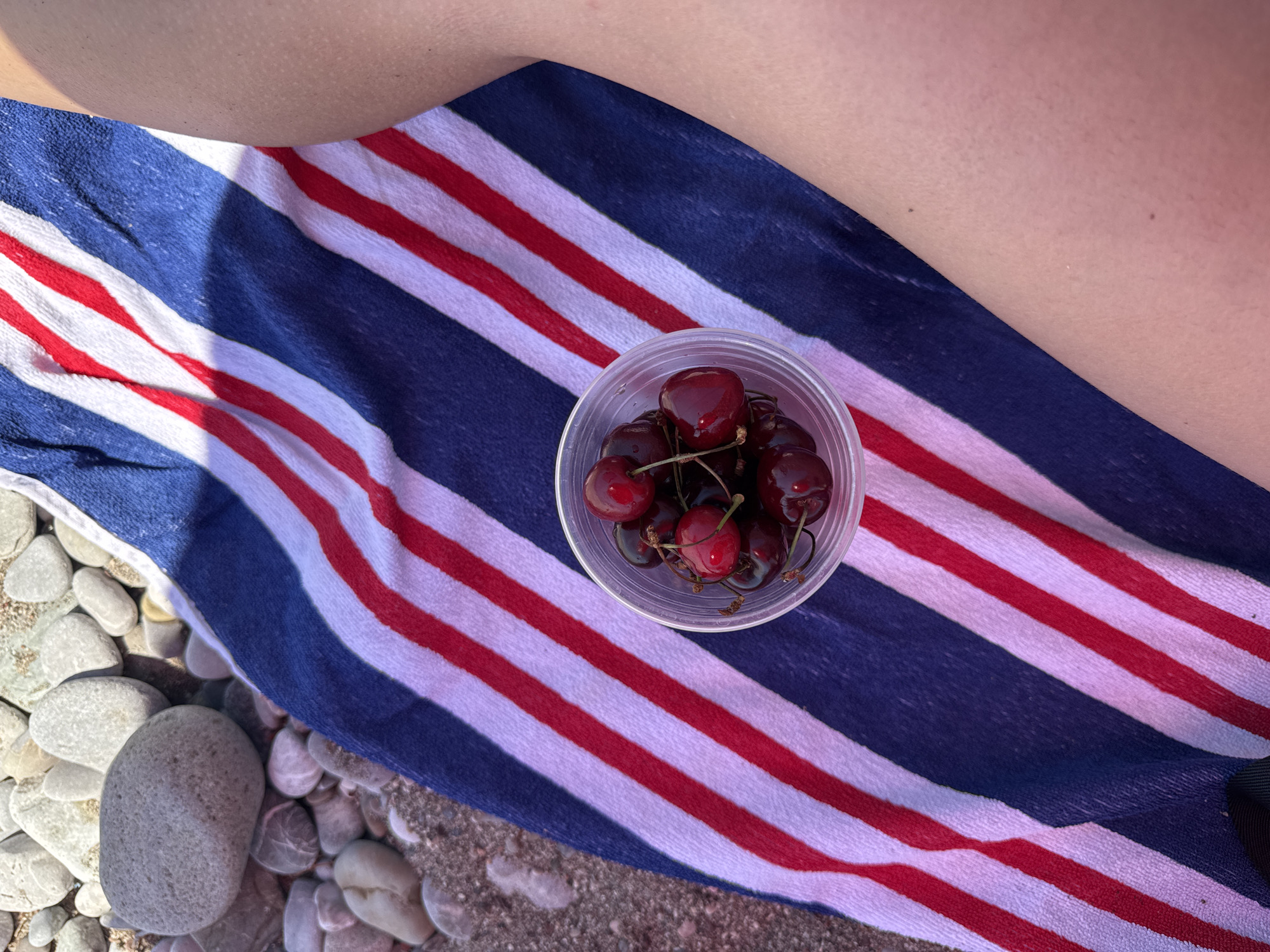 In Šušanj, Montenegro, a close-up shot captures a beach scene. A person's leg, with visible goosebumps and a light tan, rests on a striped towel featuring diagonal red, white, and blue bands. The towel is laid out on a pebble beach. To the left of the towel, a clear plastic cup filled with dark red cherries is positioned. The background consists of various smooth, grey and white pebbles, characteristic of a Mediterranean shoreline.