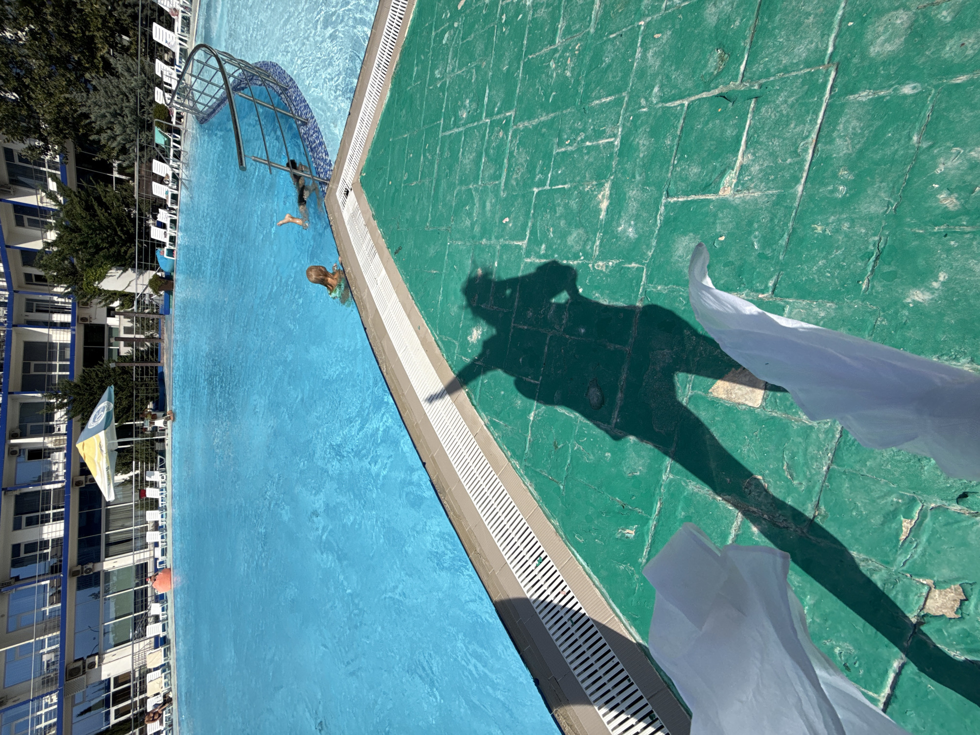 The image shows a swimming pool area with a hotel in the background. The pool is filled with bright blue water and several lounge chairs are arranged around it. There are two children swimming in the pool, one of whom is partially submerged. A shadow of a person holding a camera is cast across the green tiled ground in the foreground. White fabric is draped over the tiles. The hotel appears to be several stories tall with a modern architectural design and balconies. Trees and other foliage are visible behind the hotel. The location is identified as Sevastopol, Ukraine.