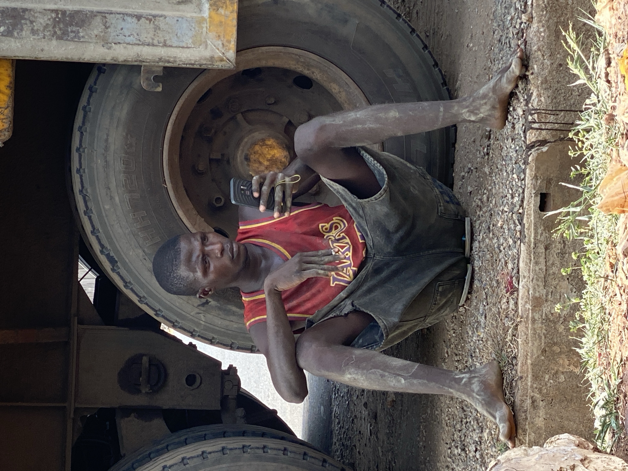 A man in a Lakers jersey and dark shorts is lying under a large truck tire in Adankolo, Nigeria. His arms and legs are dusty, and he is holding a black cell phone. The environment appears to be a roadside with vegetation present.