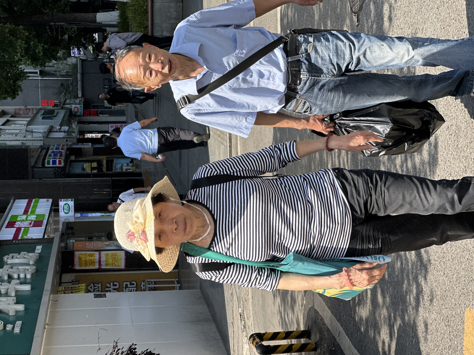 Two elderly individuals are on a sidewalk in Yuzhong District, China. The woman in the foreground wears a striped shirt, black pants, a straw hat with a flower, and carries a light green bag. The man behind her wears a light blue shirt, jeans, and a dark-colored bag over his shoulder. They appear to be handling a black plastic bag together. Shops and other pedestrians are visible in the background.
