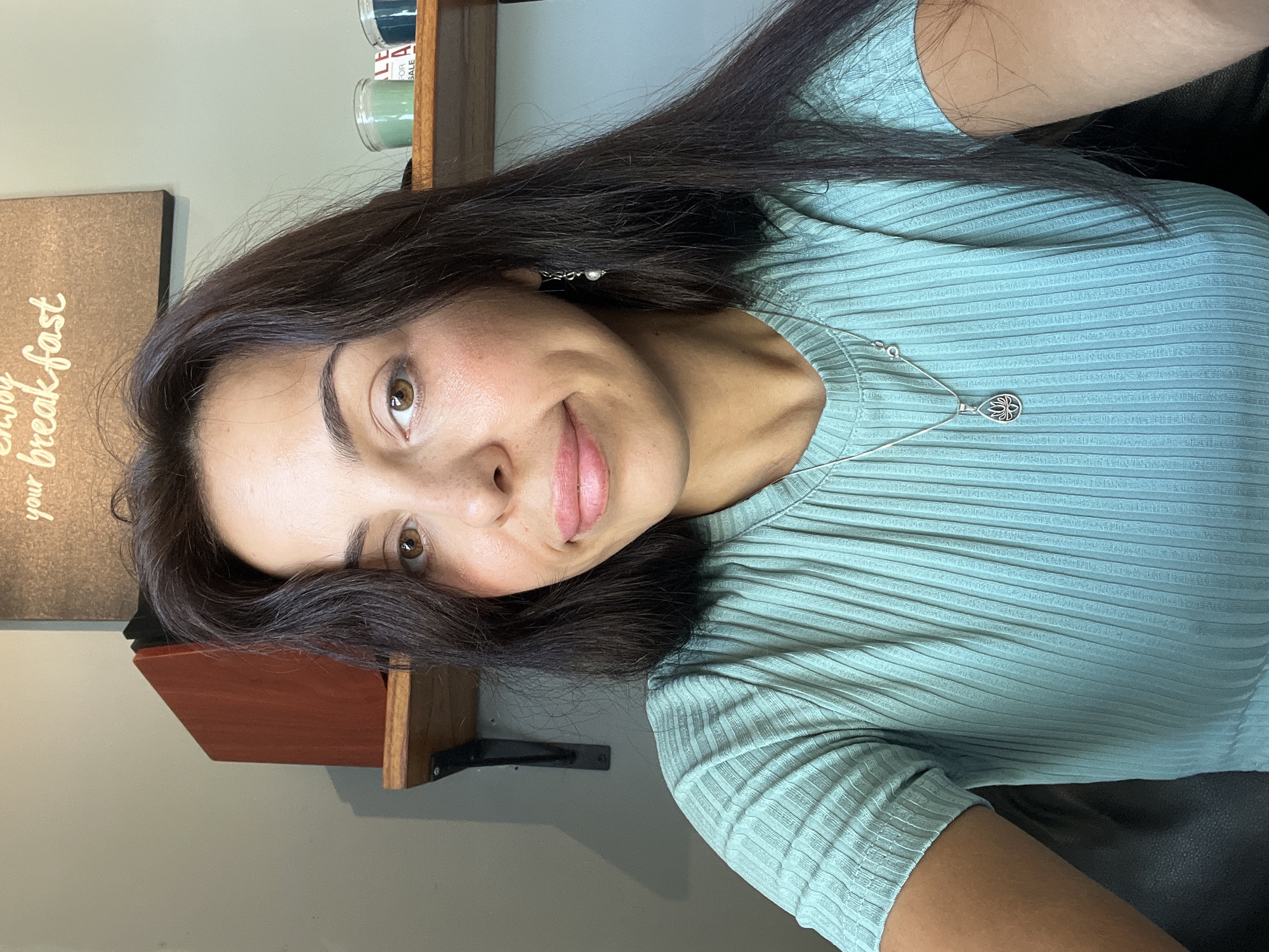 A woman with dark hair and light skin is posing for a selfie indoors. She is wearing a light green ribbed top and a silver necklace with a pendant. The background includes a wall painted in light grey, a brown wooden shelf, and a sign with the words "Enjoy your breakfast" written on it. The setting is not identifiable as Kuta Selatan, Indonesia, from the image alone.