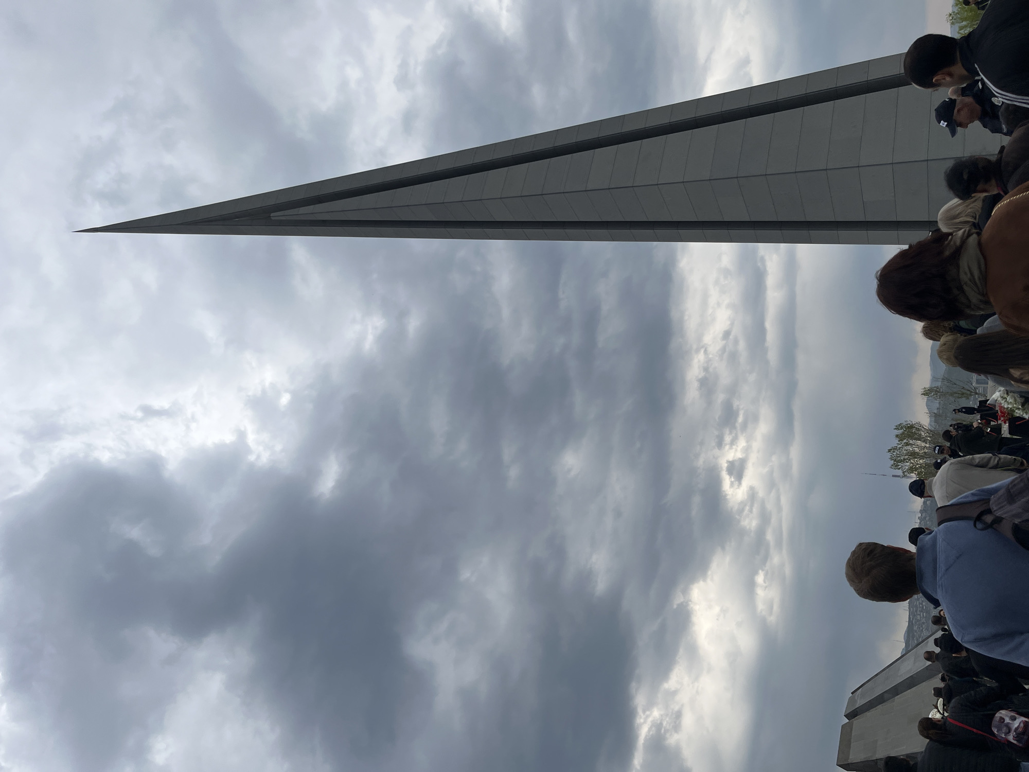 The image captures a solemn outdoor scene in Yerevan, Armenia, dominated by a towering, slender, grey concrete or stone obelisk, tapering to a sharp point at its zenith. The structure, a key element of the Armenian Genocide Memorial Complex (Tsitsernakaberd), stands against a dramatic, heavily clouded sky, with patches of lighter grey and some darker formations suggesting an overcast day.
A crowd of people is gathered in the foreground and to the right, observed mostly from behind their heads and shoulders. They appear to be looking towards the monument or beyond, suggesting a contemplative or commemorative event. Many individuals are dressed in casual attire, with some wearing hats. Their collective posture conveys a sense of respect or quiet observation. Other lower, block-like structures are partially visible in the bottom right, likely forming part of the memorial's base or surrounding architectural elements.
The weather is cloudy, with no direct sunlight visible, contributing to a subdued atmosphere. The time of day appears to be daytime, possibly late morning or afternoon, given the ambient light. There is no visible text in the image. The overall scene depicts a public gathering at a significant historical and memorial site, emphasizing its monumental scale and the solemnity of the occasion.