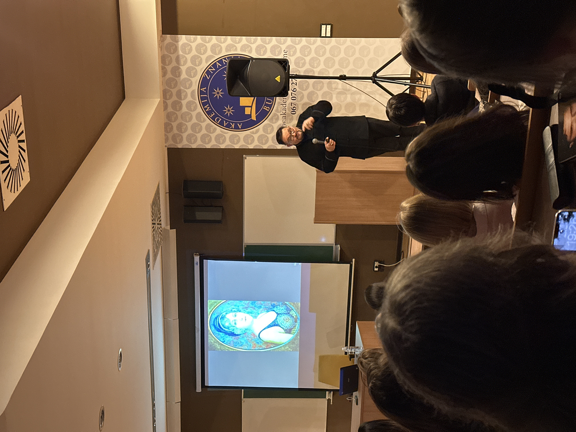 The image shows a lecture in progress. The speaker, a man in a black suit, stands on a stage addressing a seated audience. Behind him, a banner displays the emblem of "Akademija Znanja Budva," indicating this event is likely taking place at the Knowledge Academy in Budva, Montenegro. A projector displays an image of a woman on a screen. The audience consists of multiple rows of people, with the front rows more visible, suggesting they are attentively watching the presentation. The lighting in the room is moderate, illuminating the stage area but leaving the audience in more subdued light. A sound system with a microphone stand is set up near the speaker.