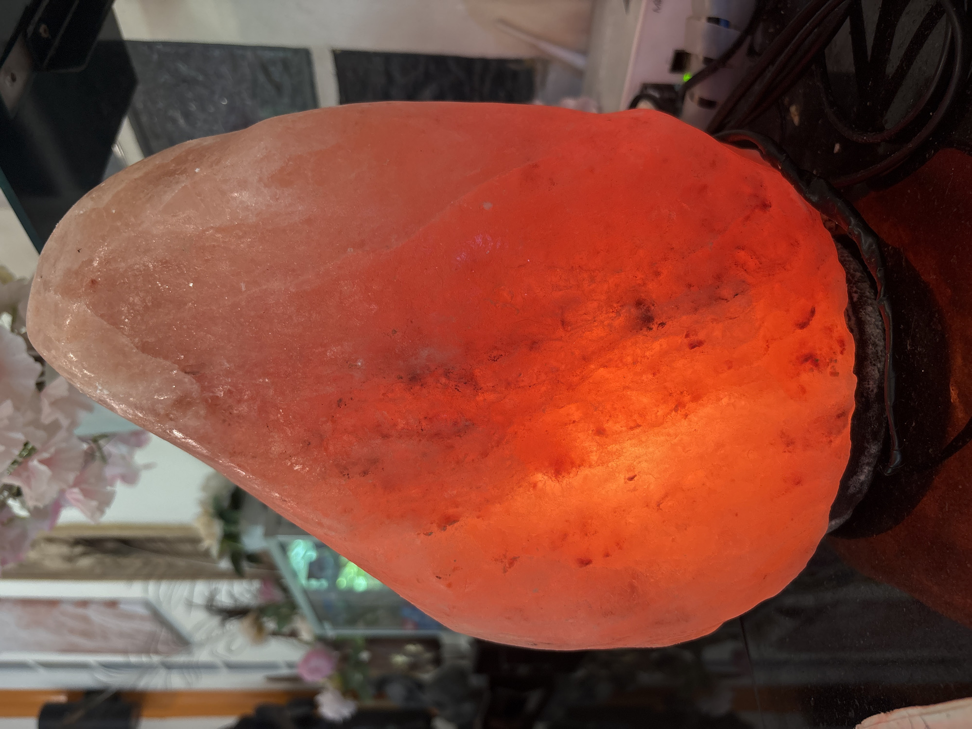 The image shows a lit Himalayan salt lamp on a black reflective surface. The lamp is irregularly shaped and glows with an orange-red light, with lighter pinkish tones towards the top. The background is out of focus, but appears to be an indoor setting with shelves containing decorative items, including flowers and possibly figurines. A power cord is visible near the base of the lamp. There are no people or animals present in the image, and no specific location in Chalong, Thailand is identifiable.