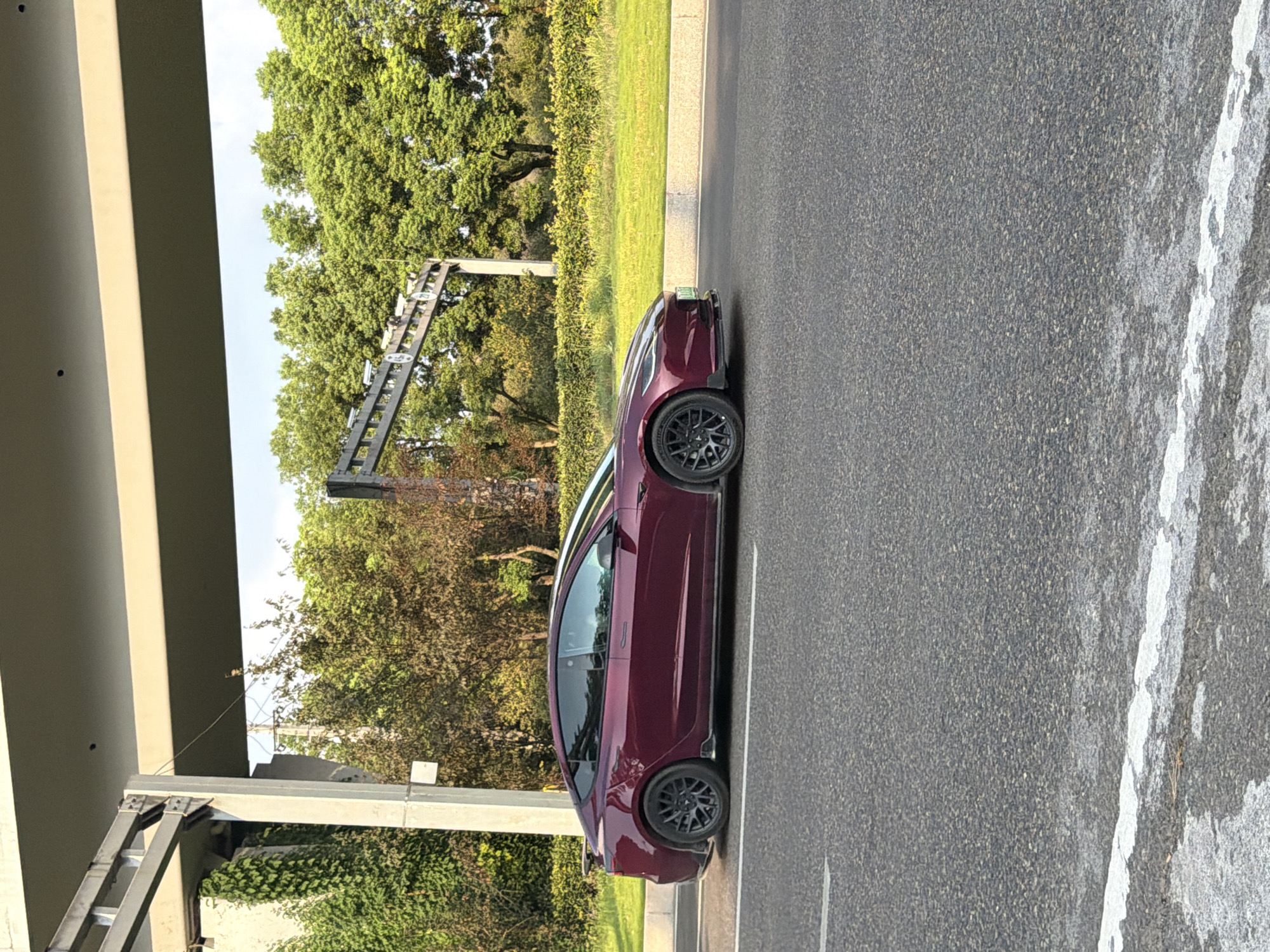 A maroon Tesla Model 3 is parked on a paved road in Jiangjin, China. The car is facing the right side of the frame and is positioned on the right lane of the road. The vehicle has tinted windows and a black spoiler on the trunk. The car is seen from its side profile. In the background, there are trees with green and brown foliage and a concrete overpass with metal beams. The sky is visible above the trees and appears to be partly cloudy. The road has white lane markings. The environmental setting suggests it is daytime and possibly late afternoon due to the lighting.