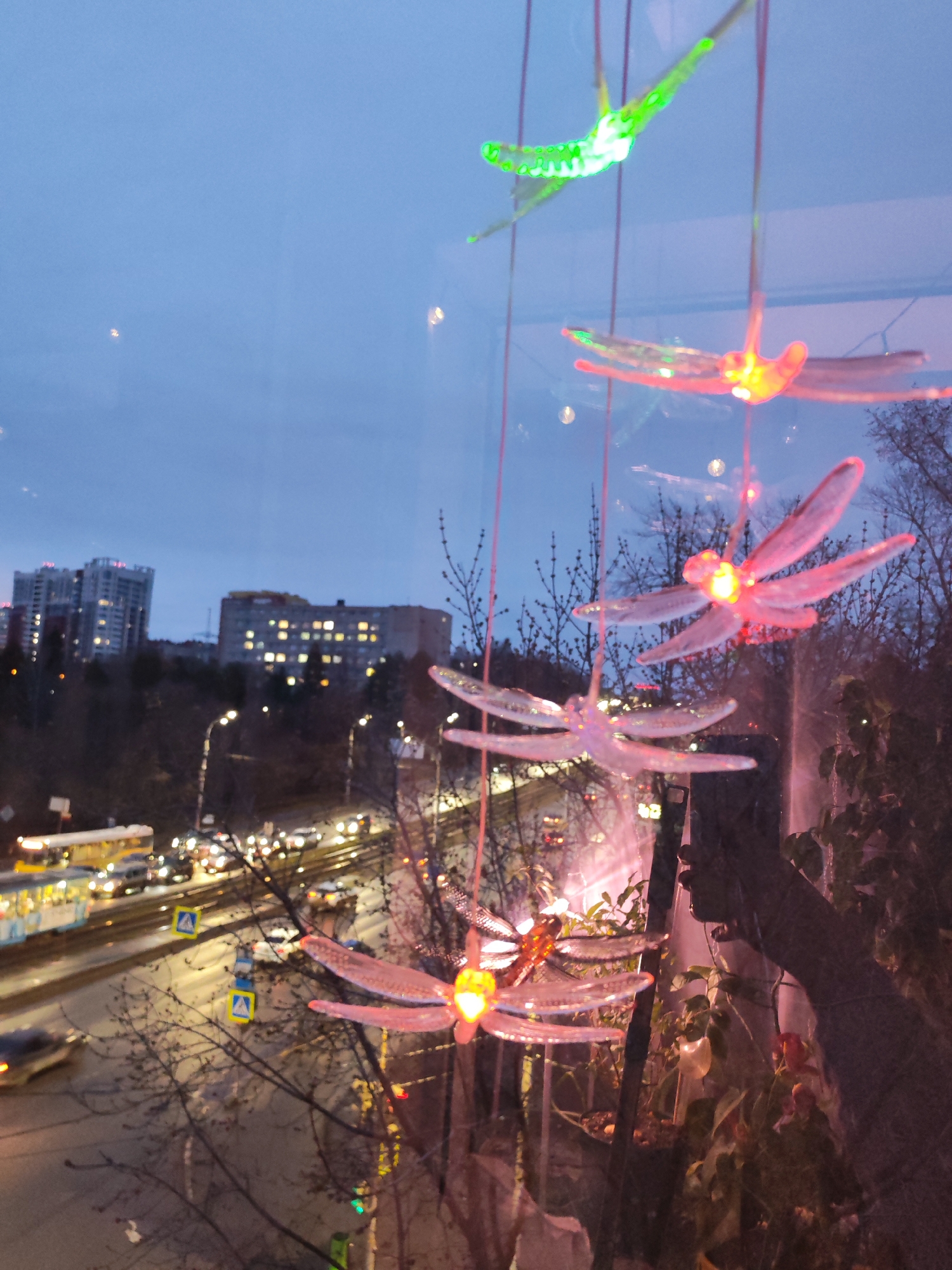 The image shows a view from a high vantage point in Izhevsk, Russia, at dusk. The main subjects are decorative, illuminated dragonflies hanging indoors, likely near a window. Through the window, a cityscape is visible, including multi-story apartment buildings with lit windows. A busy street with cars, buses, and trams is seen below, with streetlights illuminating the scene. Bare trees and some greenery fill the spaces between the buildings and the street. The overall atmosphere is a blend of urban activity and indoor decoration.