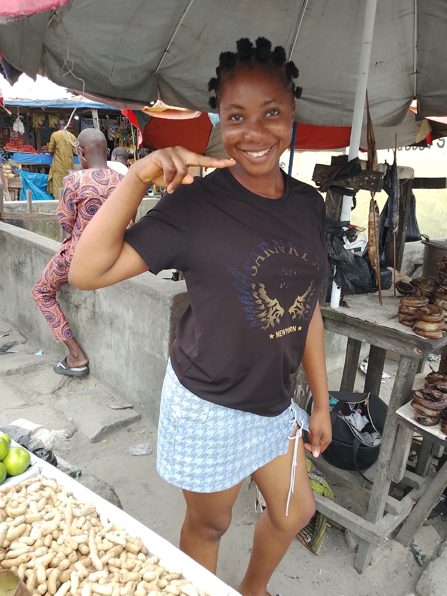 A medium shot captures a Black woman standing in a marketplace in Ado, Nigeria. She is looking at the camera and smiling. The woman has dark skin, braided hair, and is wearing a dark t-shirt and a light blue skirt. She is standing in front of a stall with fish and other goods on display. In the background, there are other stalls and people walking around. The overall scene suggests a vibrant and busy market.