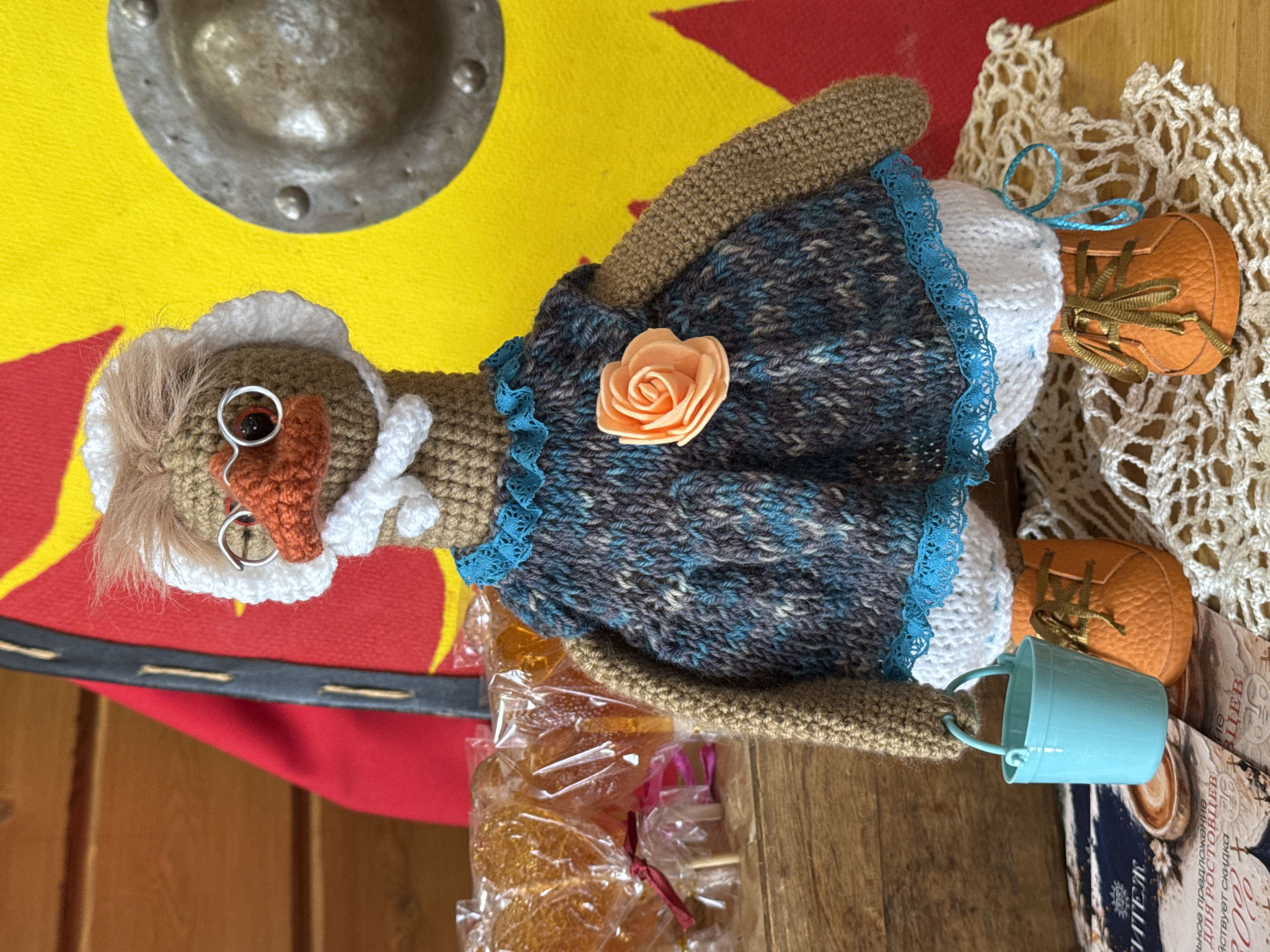 The image depicts a handcrafted doll lying on a wooden surface. The doll is dressed in a knitted outfit consisting of a blue and grey dress with a peach-colored rose embellishment and a white crocheted bonnet. The doll also has miniature orange shoes with tied laces. Beside the doll is a small blue bucket, a package of lollipops, and a piece of crocheted fabric. In the background, there's a colorful round shield with a metal center. The setting appears to be indoors, possibly a shop or craft fair. The location in the city of Unknown, Unknown cannot be determined from the image.