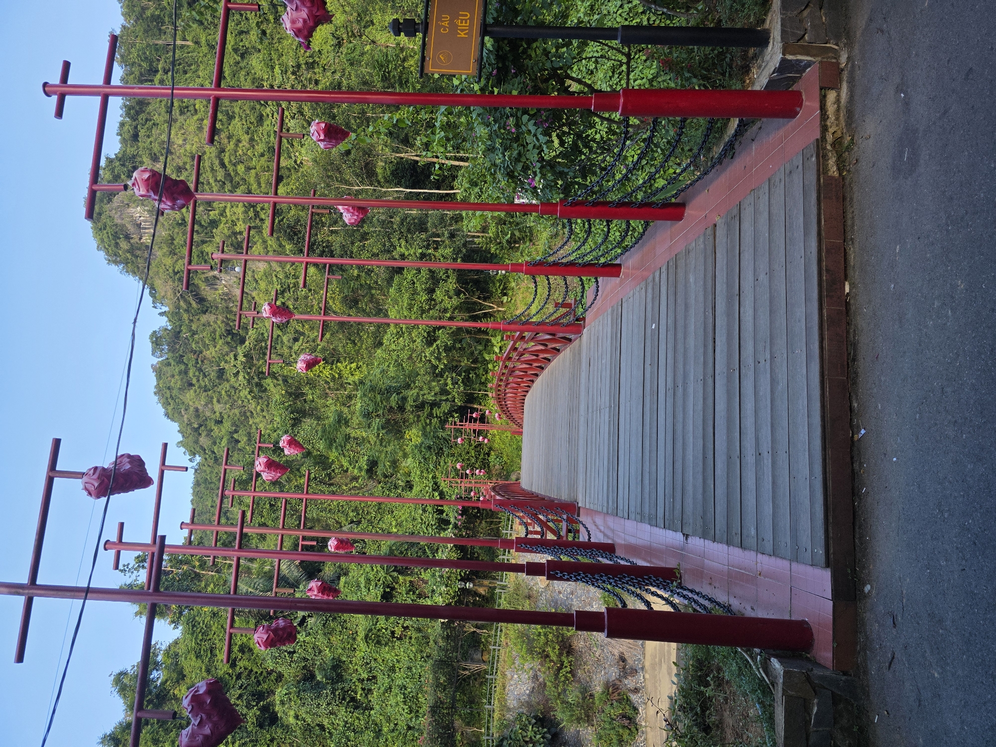 The image depicts a wooden bridge with red railings and lampposts adorned with red heart-shaped decorations. The bridge is situated in a lush, green environment with a mountainous backdrop. A signpost on the right side of the image reads "CẦU KIỀU" in Vietnamese, indicating that the bridge is located in the city of Đà Nẵng, Vietnam. The scene is captured during daylight hours with a clear sky. There are no people or animals visible in the image. The focus is on the bridge and its decorative elements, set against the natural landscape.