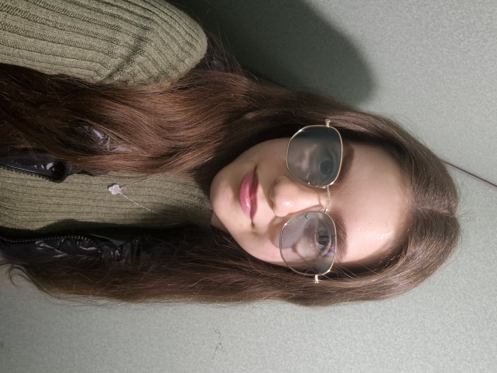 A close-up, slightly angled portrait features a young woman lying down, with the original image rotated 90 degrees clockwise. She has long, flowing reddish-brown hair that spreads out around her head. Her face is centered in the frame, and she is wearing stylish square-shaped sunglasses with reflective lenses and delicate gold-toned frames, obscuring her eyes. Her lips are painted with bright pink lipstick, showing a subtle, relaxed expression.
She is dressed in a ribbed olive green or army green sweater. Peeking out from beneath the sweater, on her left side, is a section of a black, possibly shiny, jacket or vest with a visible zipper. A delicate silver-colored necklace with a small, sparkling pendant, possibly shaped like a heart or star, rests on her sweater. The background is a plain, muted green or grey-green surface, suggesting an indoor setting such as a wall or bed. The lighting appears to be artificial, creating reflections on her sunglasses. No text is visible in the image. The photo captures a casual, posed moment indoors in Pavlikeni, Bulgaria.