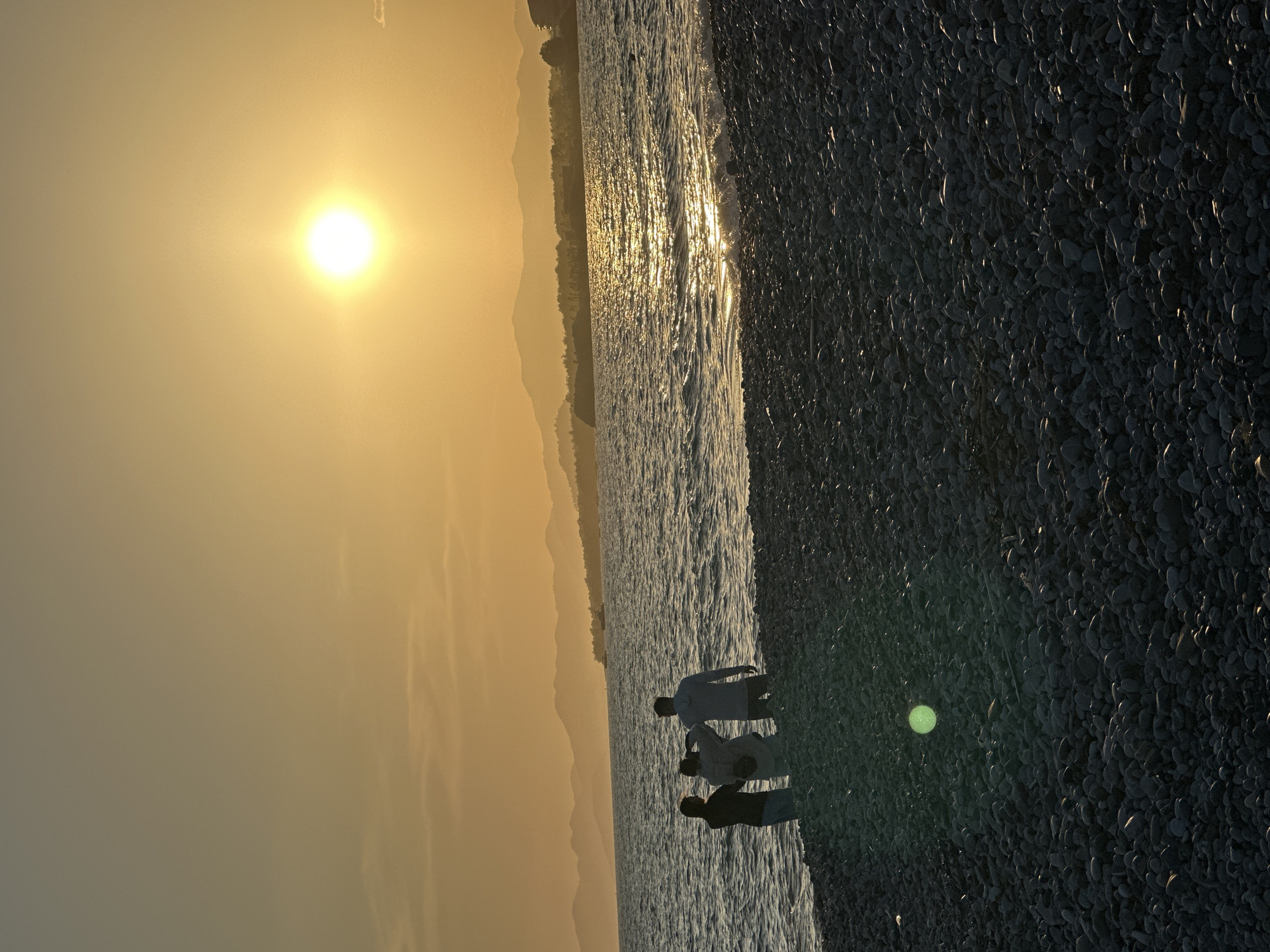 The image depicts a sunset scene at a pebble beach, likely in Fethiye, Turkey. The sun is low on the horizon, casting a warm, golden light. In the distance, there are silhouettes of mountains. The sea reflects the sunlight with a bright, shimmering effect. At the edge of the water, there are three people. The beach itself is covered in smooth, dark pebbles. A circular artifact, possibly a lens flare, is visible on the pebble beach.