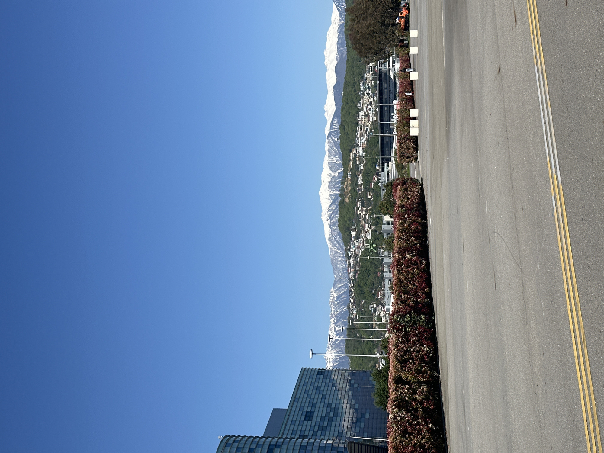 The image displays a clear, bright, and sunny day in what appears to be a developed urban or suburban area, set against a backdrop of snow-capped mountains. The scene is primarily composed of natural landscapes and modern infrastructure.
In the background, a long range of majestic mountains rises, their peaks distinctly covered in snow. Below these snow-capped summits, the mountain slopes are heavily forested with green trees, interspersed with numerous residential and commercial buildings, suggesting a populated settlement extending into the foothills.
In the mid-ground and foreground, contemporary buildings with glass facades and dark paneling are visible, reflecting the clear blue sky. A wide, paved road occupies the right side of the frame, featuring crisp double yellow lines and a single white line, indicating traffic lanes. Running parallel to the road is a dense, manicured hedge with a mix of green and reddish foliage, along with a series of light-colored, cuboid bollards or planters. Several tall, slender streetlights with circular tops are also interspersed throughout the scene.
The time of day is bright daylight, with no clouds in the vibrant blue sky, suggesting excellent weather. No people or specific activities are discernible in the image, presenting a tranquil and static view. The setting aligns with locations like Sirius, Russia, known for its mountainous terrain and modern development. There is no visible text in the image.