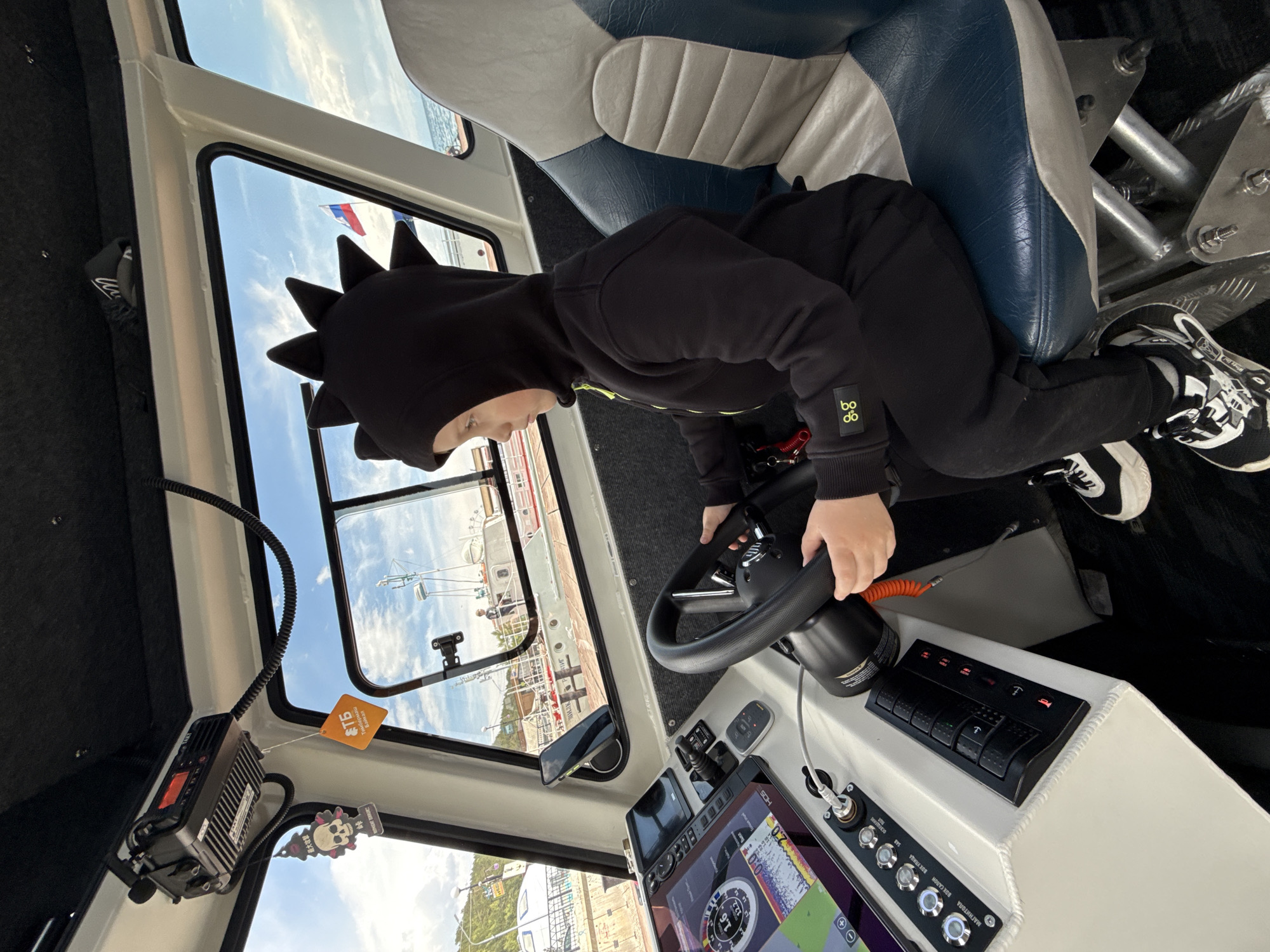 A child wearing a black hooded sweatshirt with dinosaur spikes on the hood is at the helm of a boat, steering. The child is seated in a blue and gray boat seat, hands on the steering wheel. The boat's control panel is visible, featuring a digital display showing a map, switches, and various control knobs. Through the boat's windows, a view of a harbor with boats docked along a pier can be seen. A building with a red roof is also visible on the pier. A Russian flag can be seen outside the window, likely indicating the boat is in Russia. There is a radio and other electronic equipment mounted near the window. Based on the limited background details, the location might be Listvyanka, Russia.