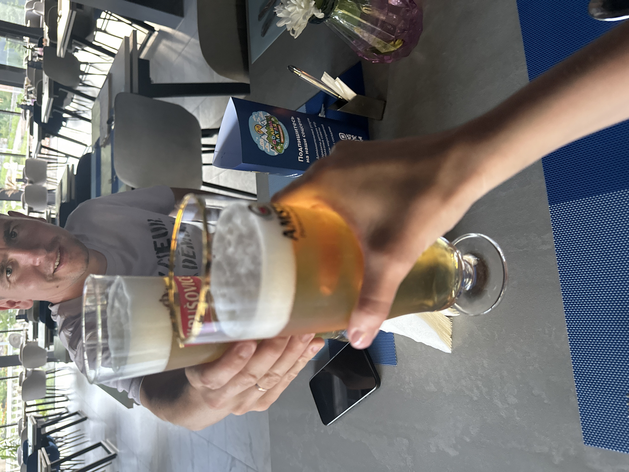 Two people are toasting with glasses of beer at a table in a restaurant. One person, a male with short hair and a light-colored t-shirt, is holding a glass in the foreground, while another person's hands are visible holding a second glass in front of him. The table is gray and set with blue placemats. A black smartphone lies on the table. In the background, there are other tables and chairs, suggesting a dining area. A blue triangular brochure with text in Russian and an illustration of mountains and a sun is visible on the table, along with a pen holder with pens and a small vase with a white flower. The scene appears to be indoors, with natural light coming from large windows, and an outdoor view of greenery visible beyond them. The location is identified as a restaurant in Danilovka (Altai Krai), Russia.