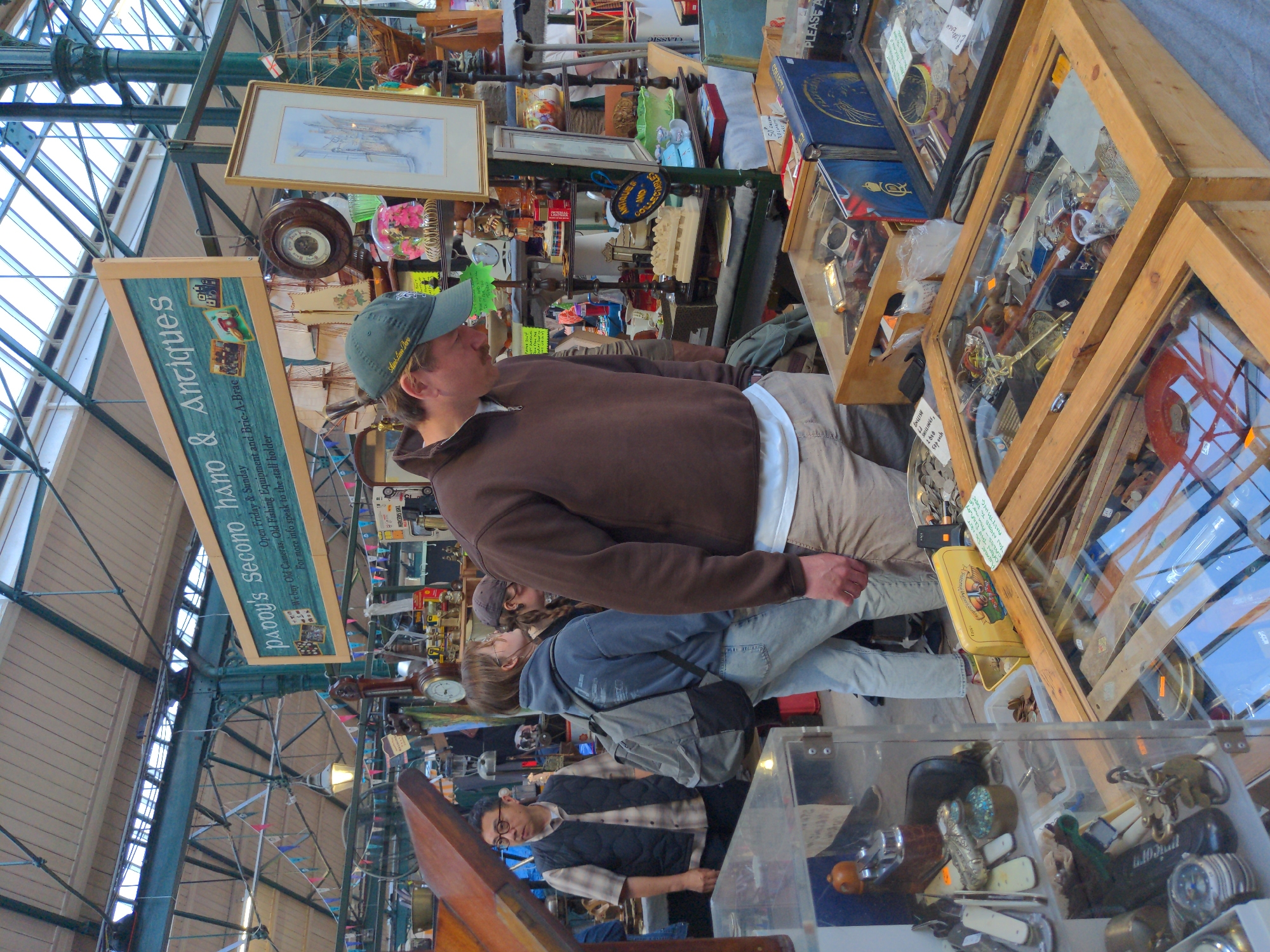 A group of people are browsing an indoor market in Belfast, United Kingdom. The market appears to be a flea market or antique market, with stalls displaying a variety of goods. The scene is crowded and bustling, with people of all ages looking through the items for sale. The market is well-lit, with natural light streaming in from a high ceiling.
In the foreground, a man wearing a green baseball cap and a brown hooded sweatshirt is examining a display of items in a glass-covered case. He appears to be a customer, and he is looking intently at the objects in the case. Behind him, a woman wearing a blue hoodie and jeans is carrying a backpack. She is looking away from the camera, and it is unclear what she is doing.
In the background, other people can be seen browsing the stalls. The stalls are filled with a variety of items, including antiques, collectibles, and clothing. The market is a lively and vibrant place, and it is clear that people are enjoying themselves.
The market is located in a large, open building with a high ceiling. The ceiling is made of glass, and it allows natural light to stream into the market. The walls of the building are made of brick, and they are decorated with a variety of signs and posters. The market is a popular destination for tourists and locals alike, and it is a great place to find unique and interesting items.