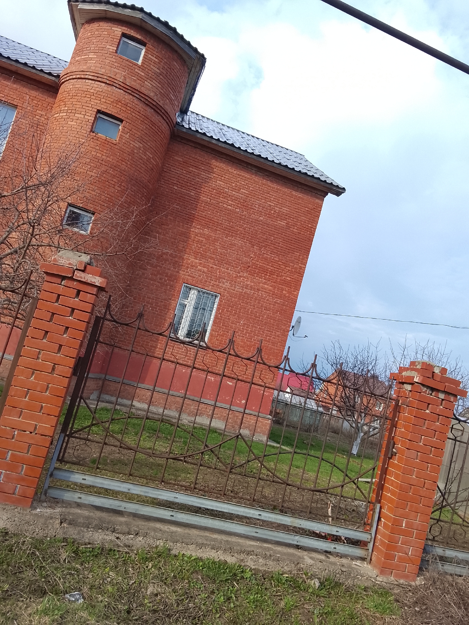 A low-angle shot captures a red brick house behind a metal fence. The house is a two-story structure with a cylindrical tower-like addition on the left side. The roof is gray and appears to be made of tiles or shingles. The tower has small windows on each floor.
The metal fence has two brick pillars marking its start and end. The fence is rusted and features a decorative pattern of curves and straight lines. The ground in front of the fence is covered in patchy grass and some dry vegetation.
In the background, behind the fence, other houses are visible, suggesting a residential area.
The image was likely taken in Chaikovsky, Russia. The presence of brick structures and residential housing aligns with the urban landscape of many Russian cities. The specific location within Chaikovsky cannot be determined without further information.