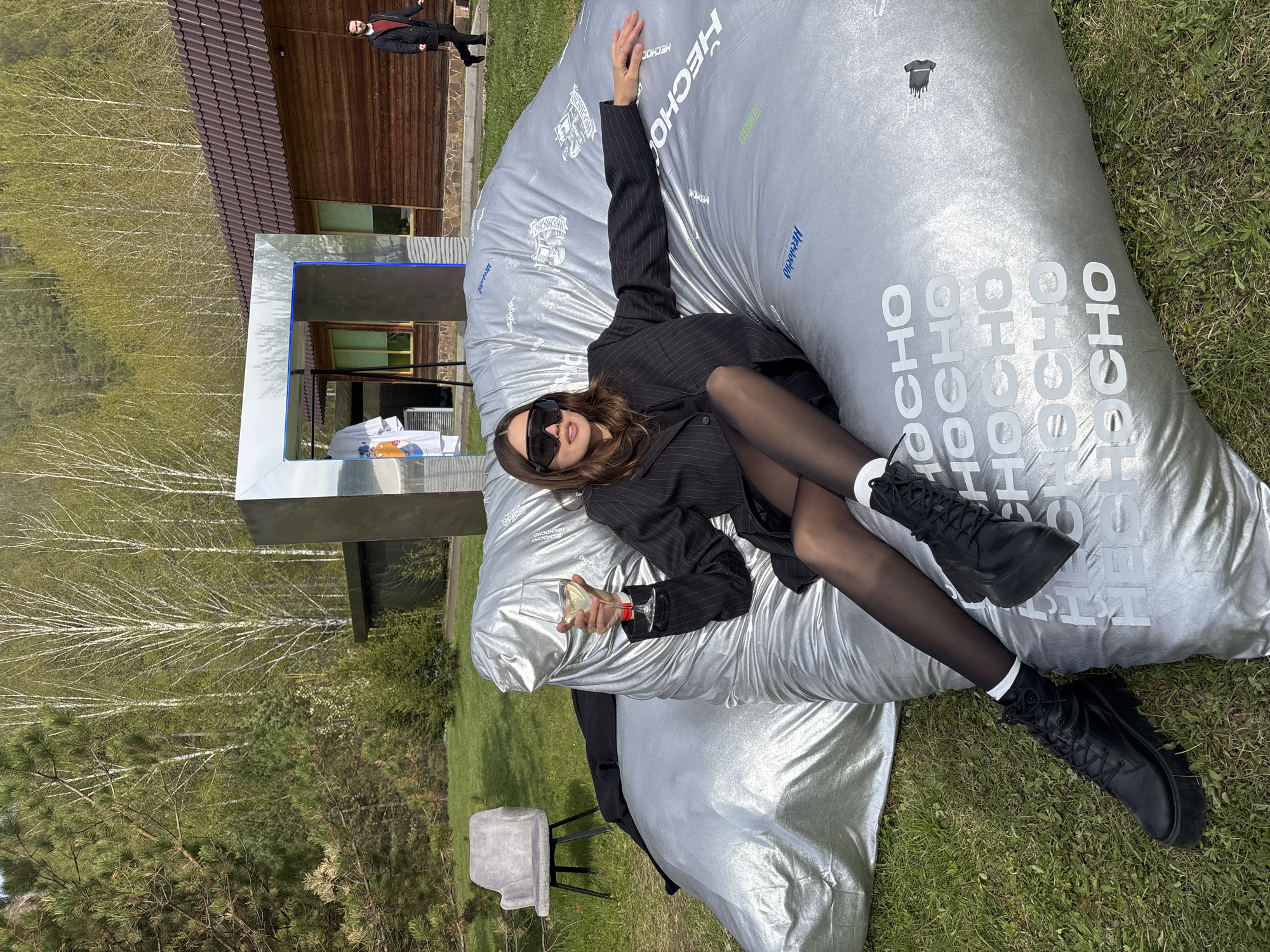 A woman is reclining on a large silver beanbag on grass. She is wearing a black pinstripe blazer, black tights, and black boots with white socks. She also wears dark sunglasses and holds a wine glass in her right hand. The beanbag has the words "HECHO" and "OCHO" printed on it. In the background, there's a building with wooden walls and a brown roof, a reflective metal structure, and a birch tree grove. The scene is set in what appears to be an outdoor area, possibly in Krasnoyarsk, Russia. A person in a black coat and red shirt is visible in the background, walking near the building. A gray chair is to the left of the beanbag.