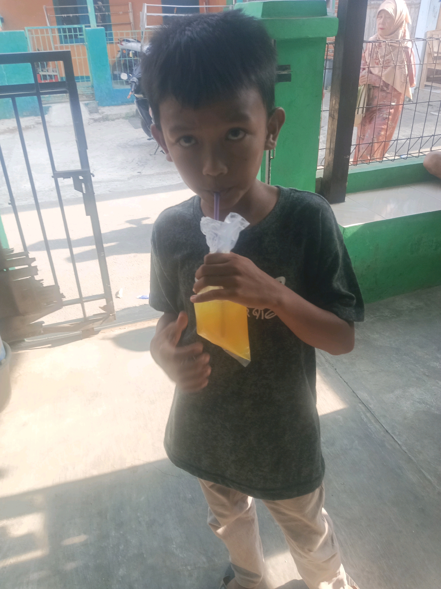 A young boy with short, dark hair is standing outdoors in Purwakarta, Indonesia, holding a clear plastic bag filled with yellow liquid and a straw. He is wearing a dark, short-sleeved t-shirt and light-colored pants. Sunlight casts shadows on the ground, suggesting it is daytime. In the background, a woman in a light-colored hijab and patterned dress is visible behind a fence. There is also a motorcycle and some buildings in the background. The environment appears to be a residential area with a paved ground surface.