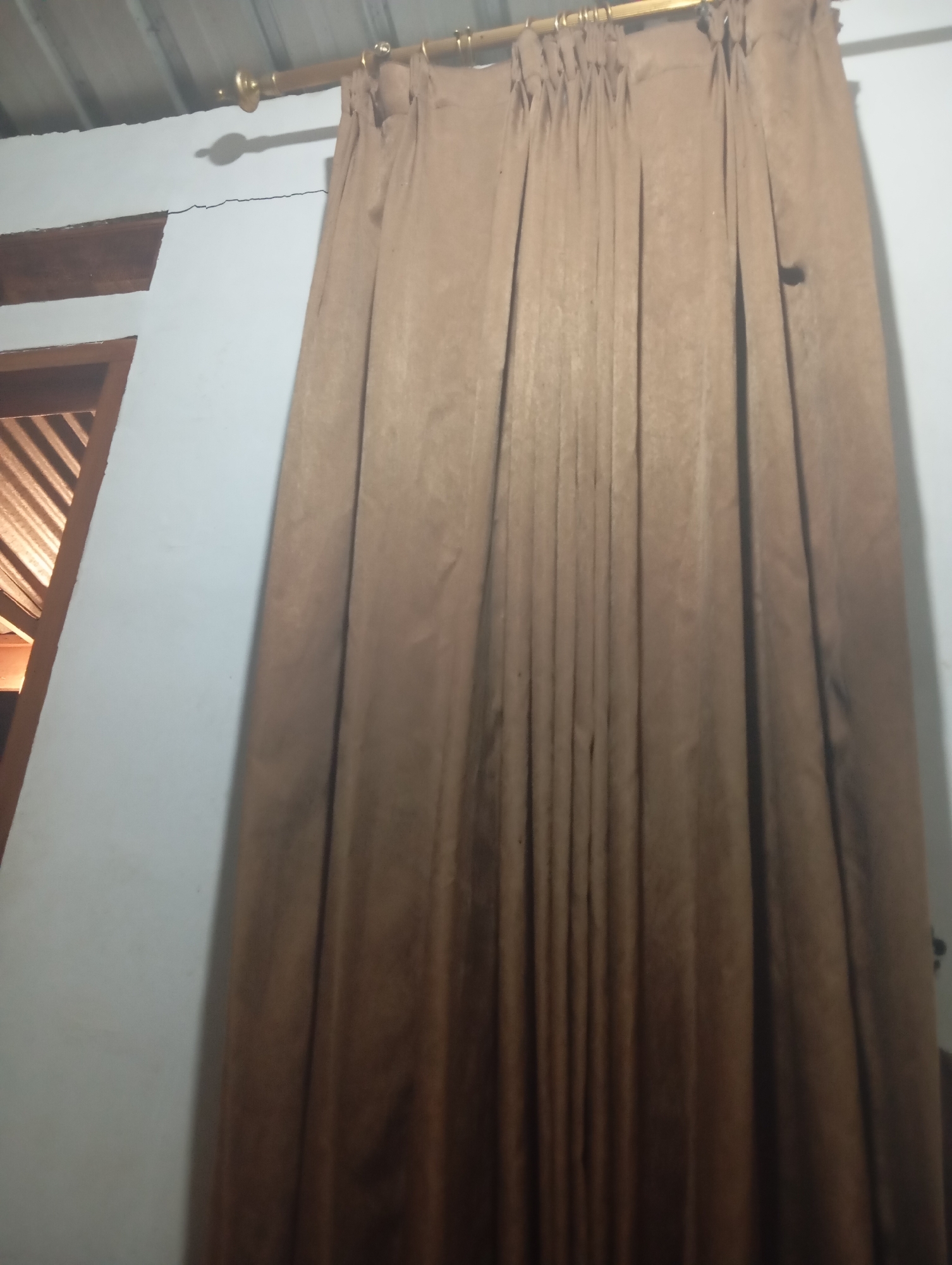 A full shot, taken from a low angle, shows a brown curtain hanging from a gold curtain rod. The curtain covers most of the frame and is drawn closed, revealing its pleats and texture. To the left of the curtain, a white wall is visible, with a wooden window frame peeking out from behind the curtain. A crack is visible in the wall above the window. The ceiling, made of corrugated metal, is visible at the top of the frame. The scene appears to be indoors.
