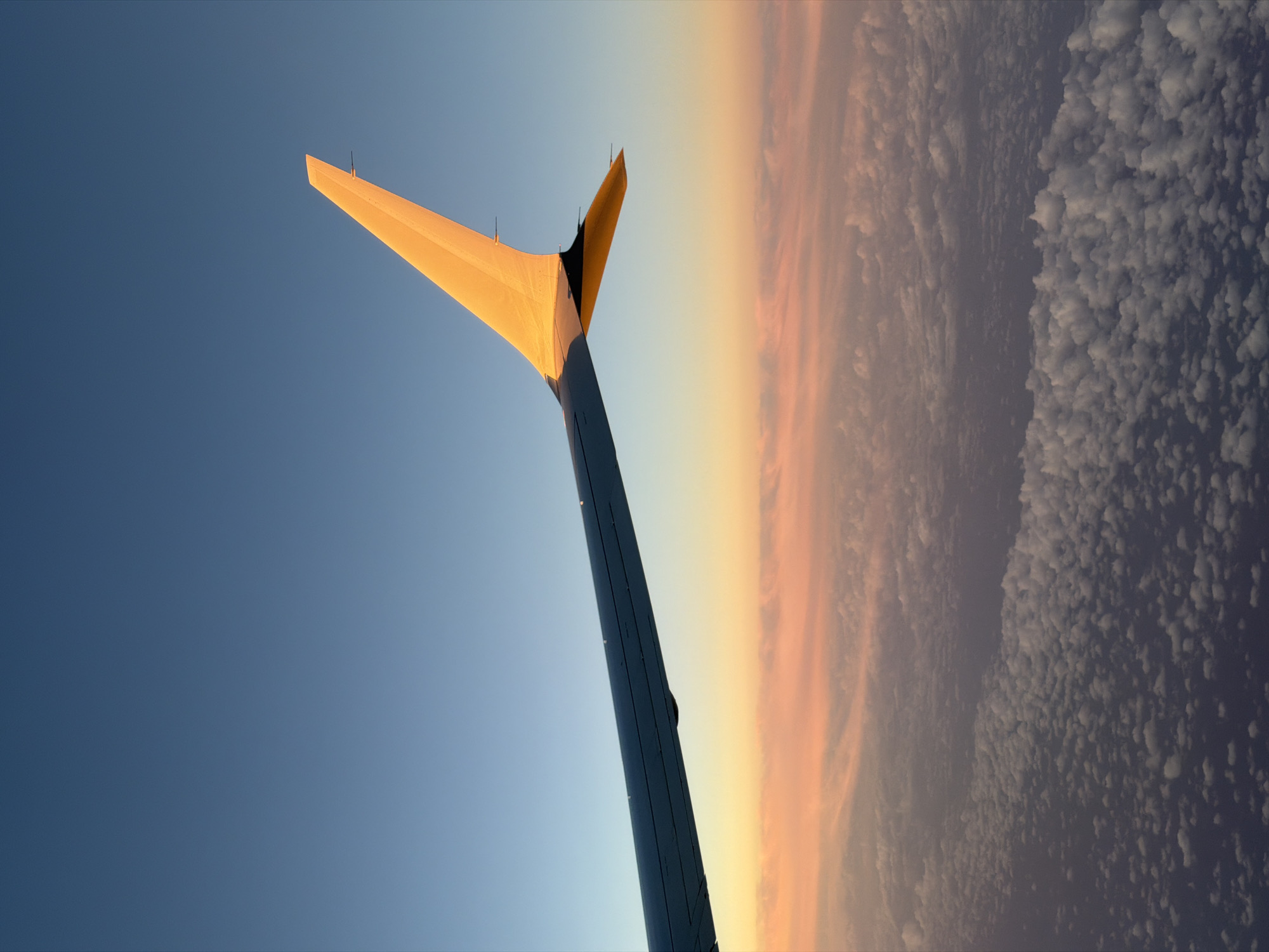 The image shows an airplane wing against a sky with a cloudscape. The wing, which is predominantly dark with a light-colored tip, extends diagonally across the frame. The sky is divided into areas of clear blue, and dense clouds, tinted with orange light. There is no information on the city, or location of Unknown, Unknown.