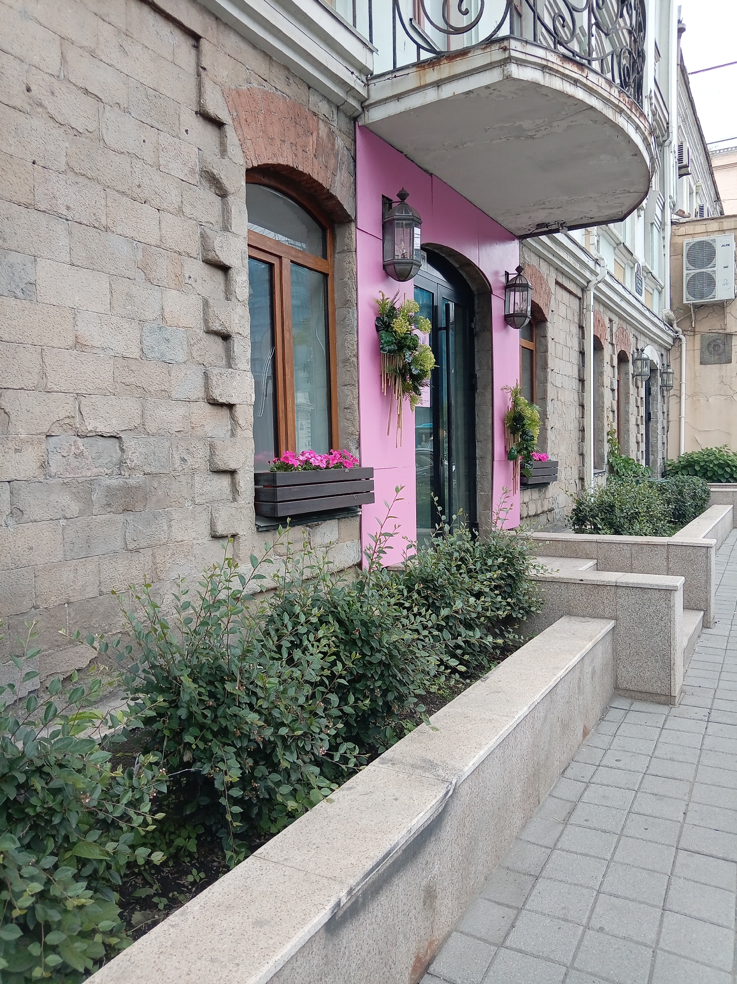 The image depicts the exterior of a building with a pink facade and stone walls. The building features arched windows and a balcony with ornate metal railings. Plants and flowers are visible in window boxes and in planters along the sidewalk. A paved walkway with square tiles runs along the side of the building. The scene suggests a commercial or residential property in a city. The location is identified as Vladivostok, Russia.