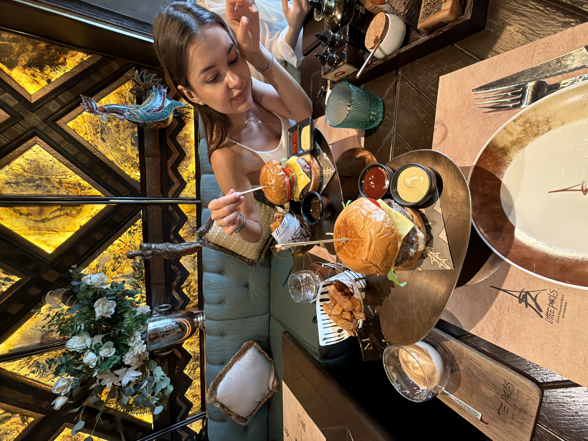 A young woman sits at a table in what appears to be a restaurant. She is holding a skewer over a hamburger. On the table are other burgers, fries, sauces, a drink, and a plate setting. The restaurant's name, "LITTLE PARIS", is visible on a wooden coaster and placemat. The restaurant's interior features ornate designs with flowers and animal sculptures. The location is likely in Choeng Thale, Thailand.