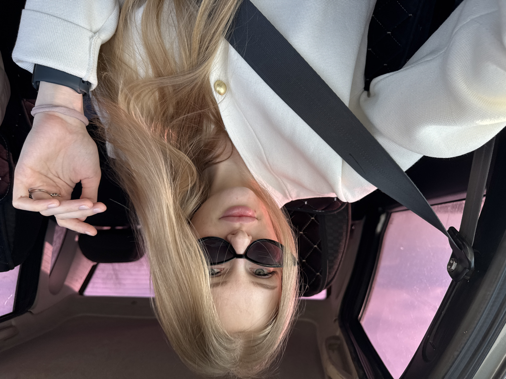 A blonde female individual is seated in the passenger seat of a vehicle in Кондратово, Russia. She is wearing a white, textured long-sleeved top with a gold button on the right chest area and black rectangular sunglasses. A black seatbelt diagonally crosses her torso. Her right hand is slightly raised, displaying a ring with small stones on her ring finger. Her left wrist, partially visible, has a dark-colored smartwatch with a matching band and a pink hair tie. The vehicle's interior includes a dark, possibly quilted, seatback and a light-colored headliner with a visible sunroof. Through the vehicle's windows, a distinct pink-purple sky is visible, with faint, bare tree branches in the distant background. The individual is looking directly towards the camera.