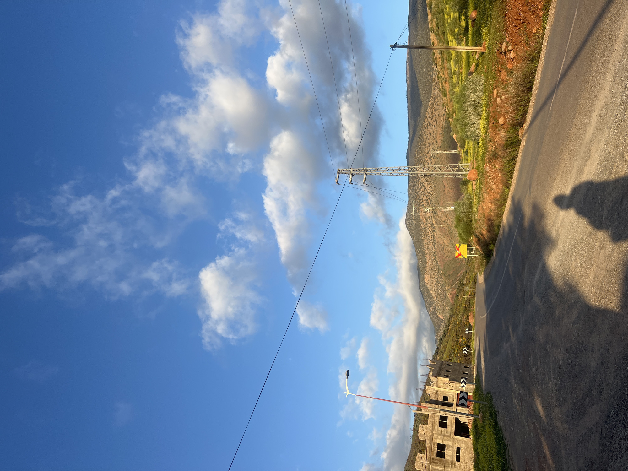 The image shows a road in Ouaouizert, Morocco, running alongside a hillside. The sky is blue with scattered clouds. Electrical power lines run across the frame. An unfinished building is visible near the road. A shadow of a person is cast onto the road surface. Vegetation covers the hillside. Road signs are present.