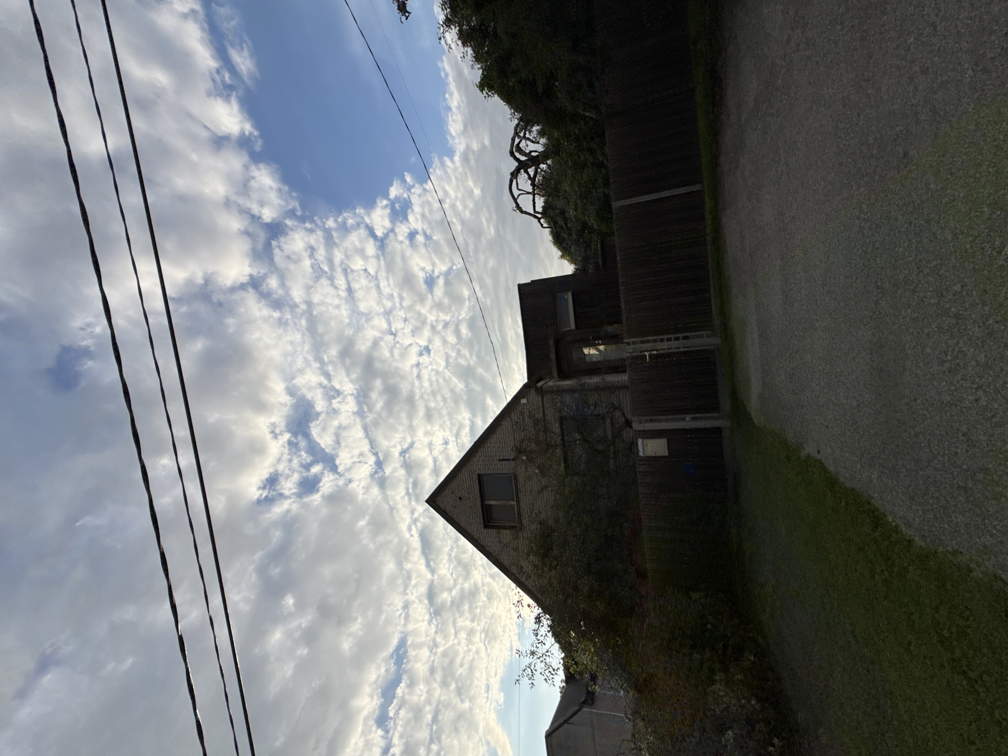 The image depicts a residential property on a cloudy day. In the foreground, a gravel driveway leads towards a wooden fence. To the left of the driveway is a patch of green grass. Behind the fence is a two-story brick house with a pitched roof and a single window visible on the upper level. Adjacent to the house is a smaller, darker structure with a glass door. Lush green trees and bushes surround the property. Overhead, three black electrical wires stretch across the frame. The sky is a mix of blue patches and scattered white clouds, with some appearing to be thin and cirrus-like. The overall scene suggests a suburban or semi-rural environment. The location of this property is in Tallinn, Estonia.