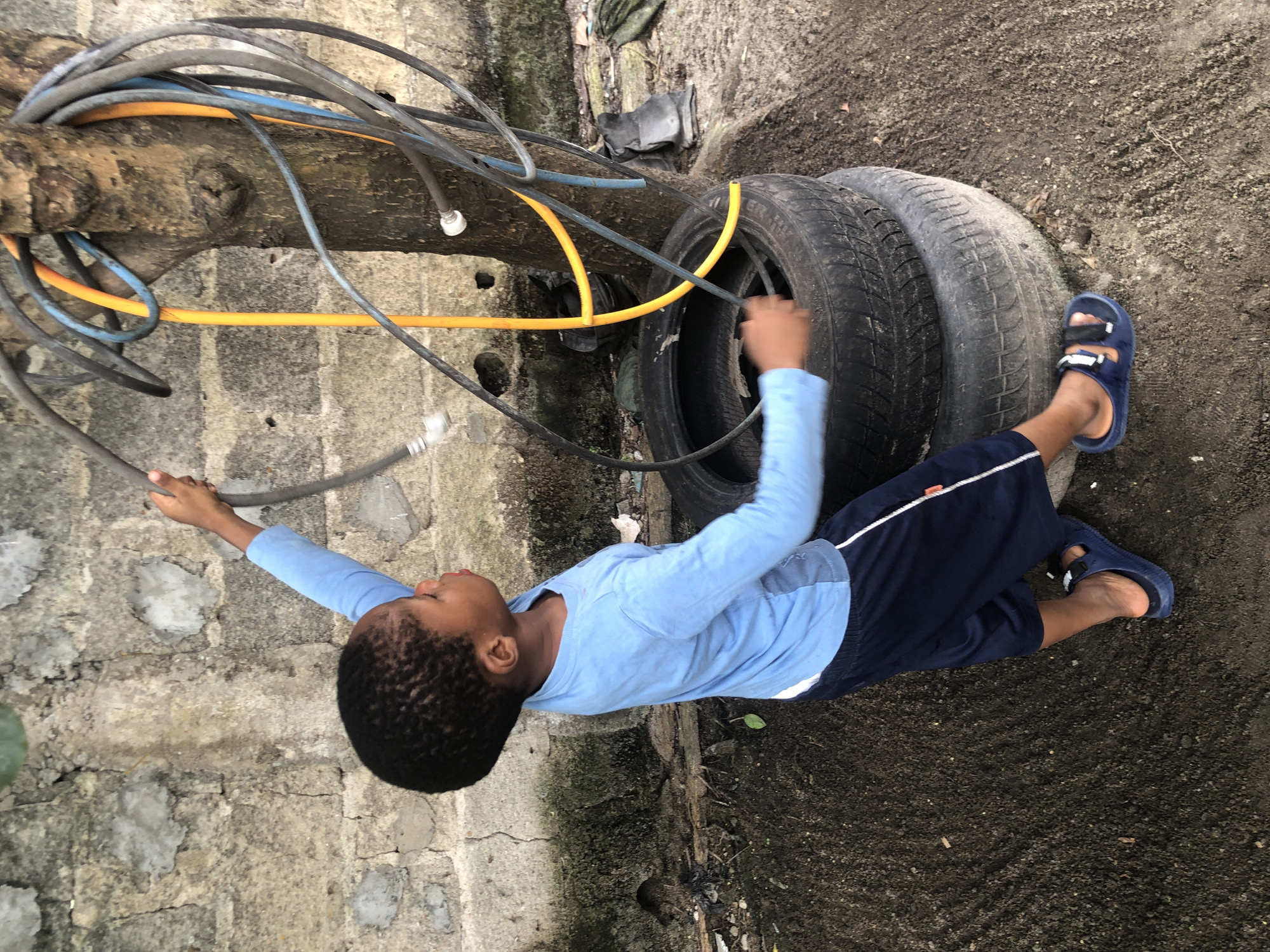 A young boy, appearing to be of African descent, is shown from the chest down, standing and holding a gray hose. He is wearing a long-sleeved light blue shirt and dark blue shorts with white trim. His feet are clad in blue sandals. The boy is positioned in front of a rough, stone wall and to the right of a tree. Several hoses of various colors, including yellow, blue, and gray, are wrapped around the tree. In front of the boy, two stacked tires lie on the ground, which is covered in dirt and debris. The overall environment suggests an outdoor, possibly informal, setting in a location that could be Ado, Nigeria, based on the provided context. The boy's posture indicates he is engaged in an activity with the hose, possibly watering or playing.