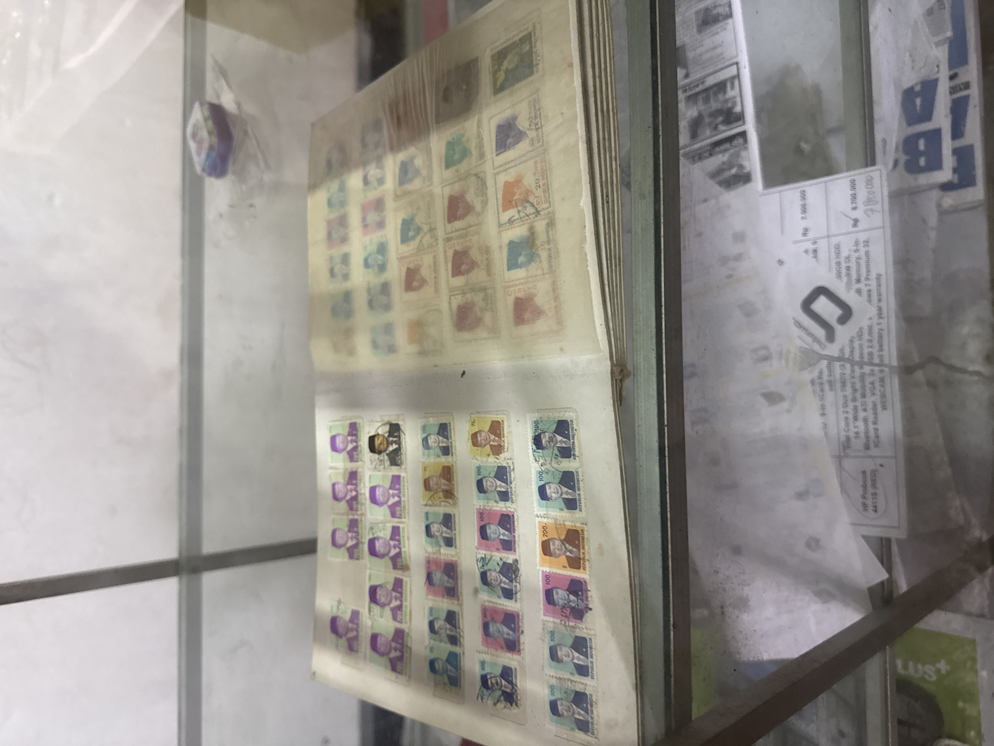 An open stamp album is displayed in a glass case. The album pages are filled with rows of postage stamps, many featuring portraits of men in formal attire. Some stamps are labeled with denominations such as "100" and "200" and text that appears to be "REPUBLIK INDONESIA". The album is positioned on a shelf within the display case. In the background, various items are visible on lower shelves, including what appears to be electronic product packaging with pricing information and a small purple box on the top shelf. The lighting suggests an indoor setting, possibly a shop or market stall, in Denpasar, Indonesia.