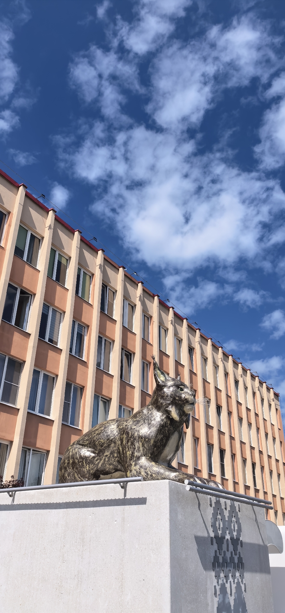 The image shows a statue of a lynx in front of a peach-colored, multi-story building with a red roofline. The building has a repeating pattern of windows with white frames. The sky is a bright blue with scattered white clouds. The lynx statue is metallic, with a textured surface that mimics fur, and is depicted in a seated position, looking to the right. The statue is positioned on a light-colored concrete pedestal that has a decorative, geometric pattern on its side. The overall scene suggests an outdoor, urban setting on a clear day. The time of day appears to be daytime, indicated by the bright sunlight and blue sky. There is no visible text in the image. The location context is Homyel, Belarus.