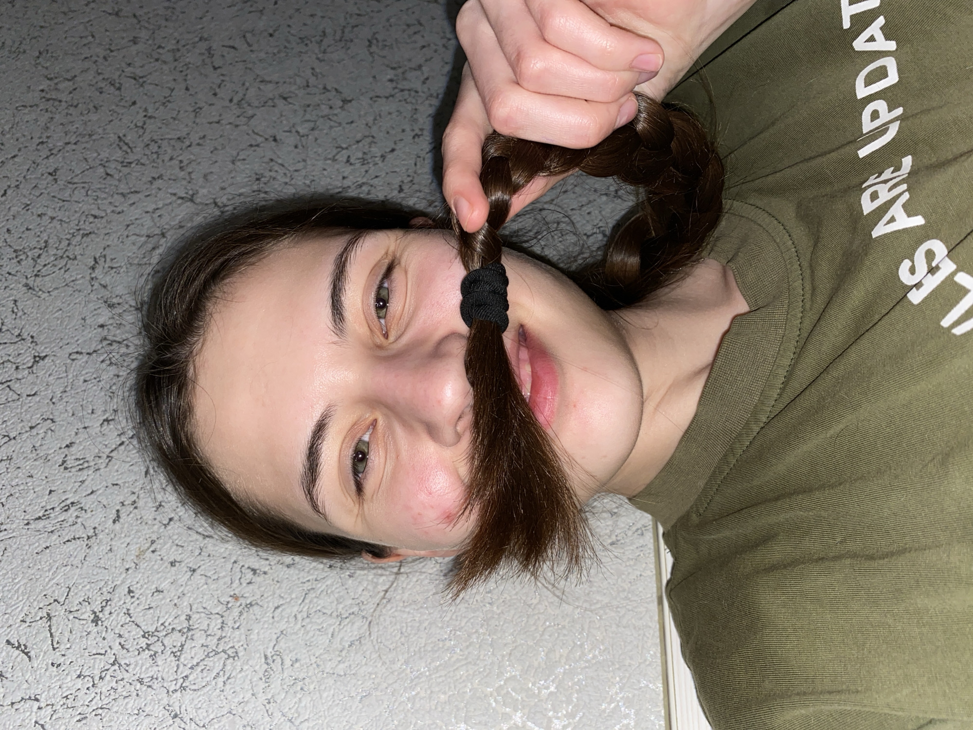 A woman lies on a grey and white textured surface, possibly a floor or countertop. She wears an olive-green t-shirt with white lettering that is partially obscured. She is holding a section of her own brown, braided hair under her nose to resemble a mustache. Her gaze is directed toward the camera. The scene is well-lit and appears to be indoors. There is no indication of the location being Votkinsk, Russia.