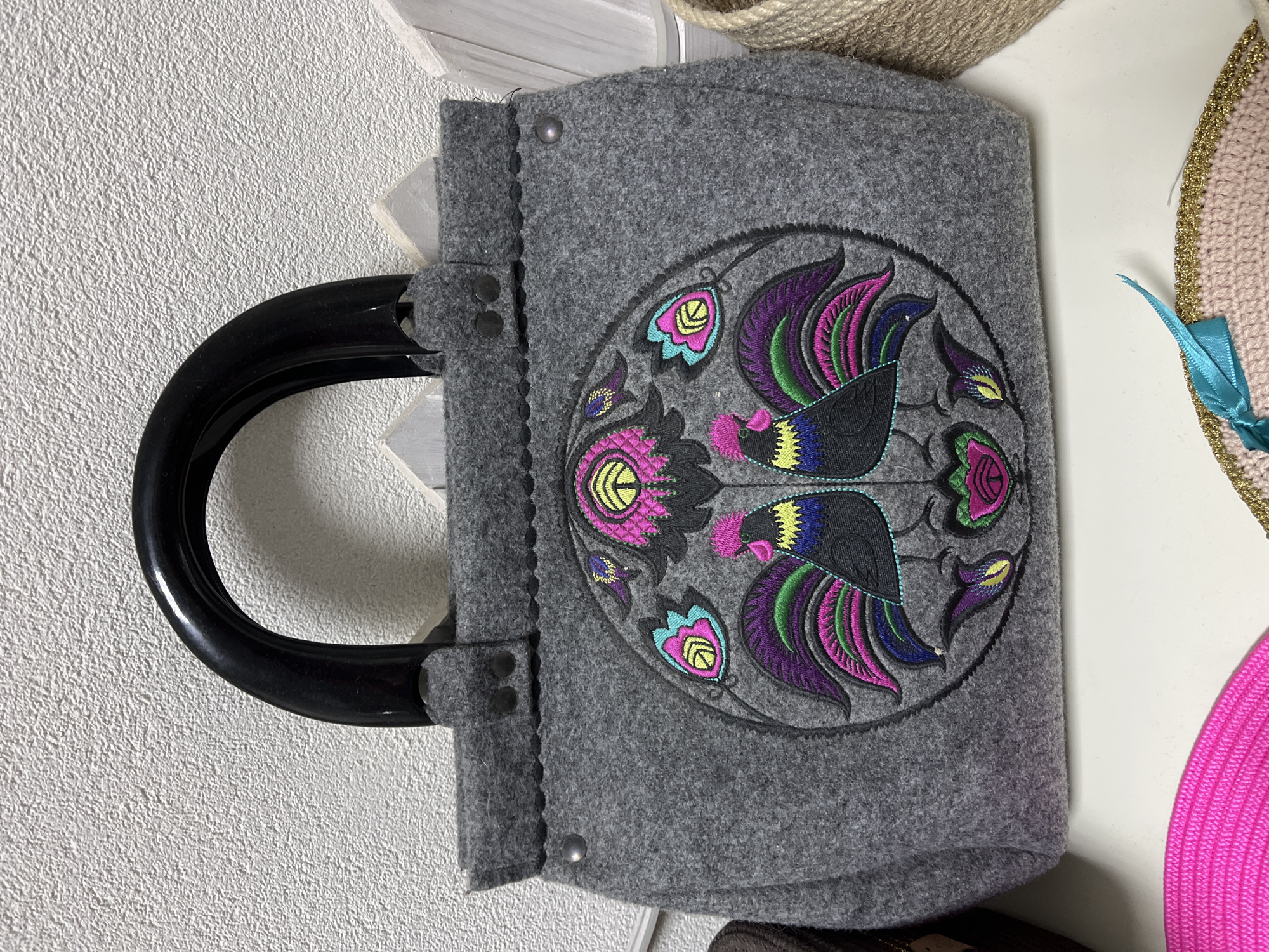 A gray felt tote bag with a black plastic handle is centered in the image. The bag features an embroidered circular design with two colorful roosters facing each other, surrounded by floral motifs in pink, purple, green, yellow, and teal. The bag is on a white surface, possibly a shelf, with a textured white wall in the background. In the foreground, a portion of a bright pink woven item and a pink crocheted item with a teal ribbon are visible. The image is a close-up of the bag and does not provide any contextual information about its location in Hrodna, Belarus.