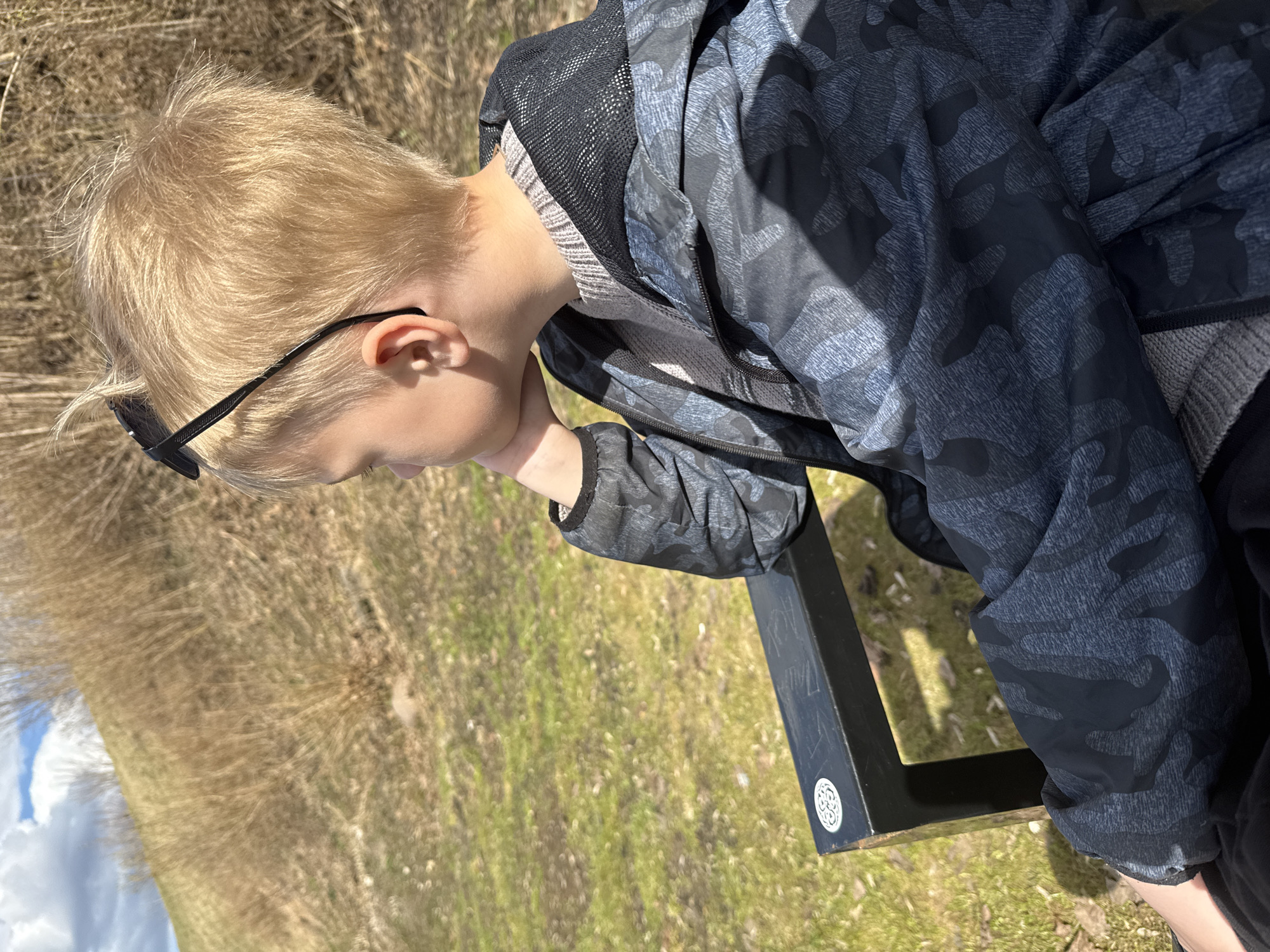A blond-haired boy wearing sunglasses is seen in an outdoor setting. He has on a gray sweater and a gray and black camouflage patterned jacket. He is resting his chin on his hand and looking down. The background includes grass, bare trees, and a partial view of a black bench. There is no indication of the location being Dmitrov, Russia.