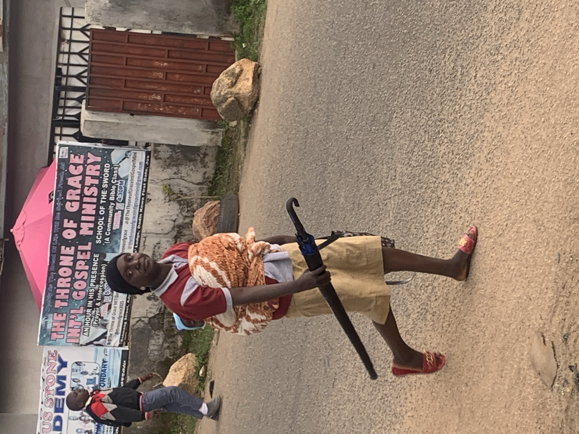 In Jos, Nigeria, a woman walks with a child strapped to her back, holding a black umbrella. She wears a red and white shirt, a yellow skirt, and red slippers. Behind her, a banner announces "THE THRONE OF GRACE INT'L GOSPEL MINISTRY" and "SCHOOL OF THE SWORD." Another banner in the background advertises "OUS STONE ACADEMY." A young boy in a jacket and jeans walks in the opposite direction. Rocks and overgrown vegetation are visible along the roadside.