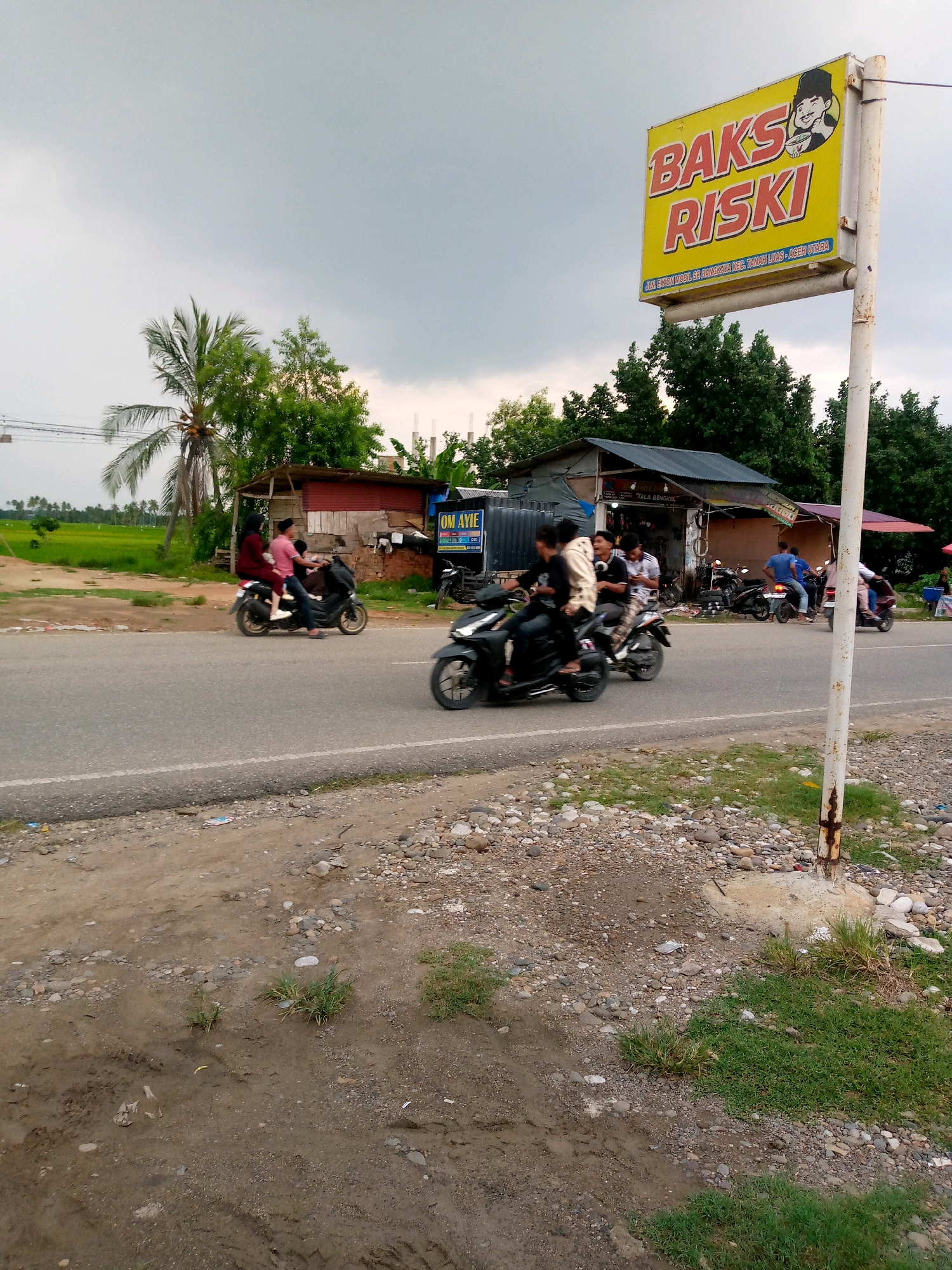 In Gampongpaya, Indonesia, a roadside scene unfolds under an overcast sky. A yellow signpost with red lettering reading "BAKS RISKI" stands prominently on the right, featuring a cartoon drawing of a man holding a bowl of food. Adjacent to the sign, a building with a corrugated metal roof and a red overhang is visible, with several motorcycles parked in front.
The road is paved and has a white dividing line. Several people are on motorcycles, actively navigating the street. On the left, two people on a black motorcycle are traveling away from the viewer. Further down the road, a group of three people ride on a black motorcycle, two seated and one standing behind. Another motorcycle with two riders is further on. On the right side of the road, two individuals are on a motorcycle, and another motorcycle is parked.
The environment includes a grassy field and palm trees on the left side of the road, with more trees and a few buildings in the background. The foreground features a dirt and gravel embankment bordering the road.